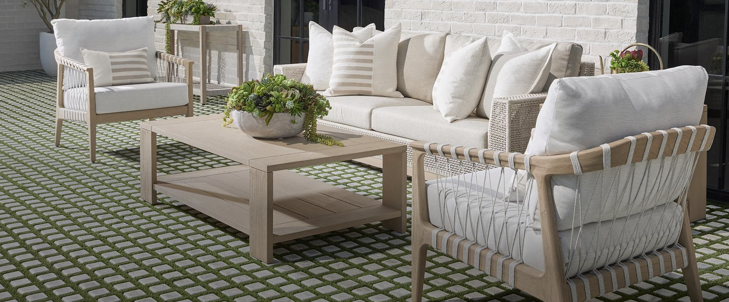 Outside patio furniture set with a beige cushioned sofa, two beige cushioned armchairs with wooden frames, and a wooden coffee table with a decorative bowl of plants, on a patterned patio with grass-filled concrete pavers, against a brick wall.