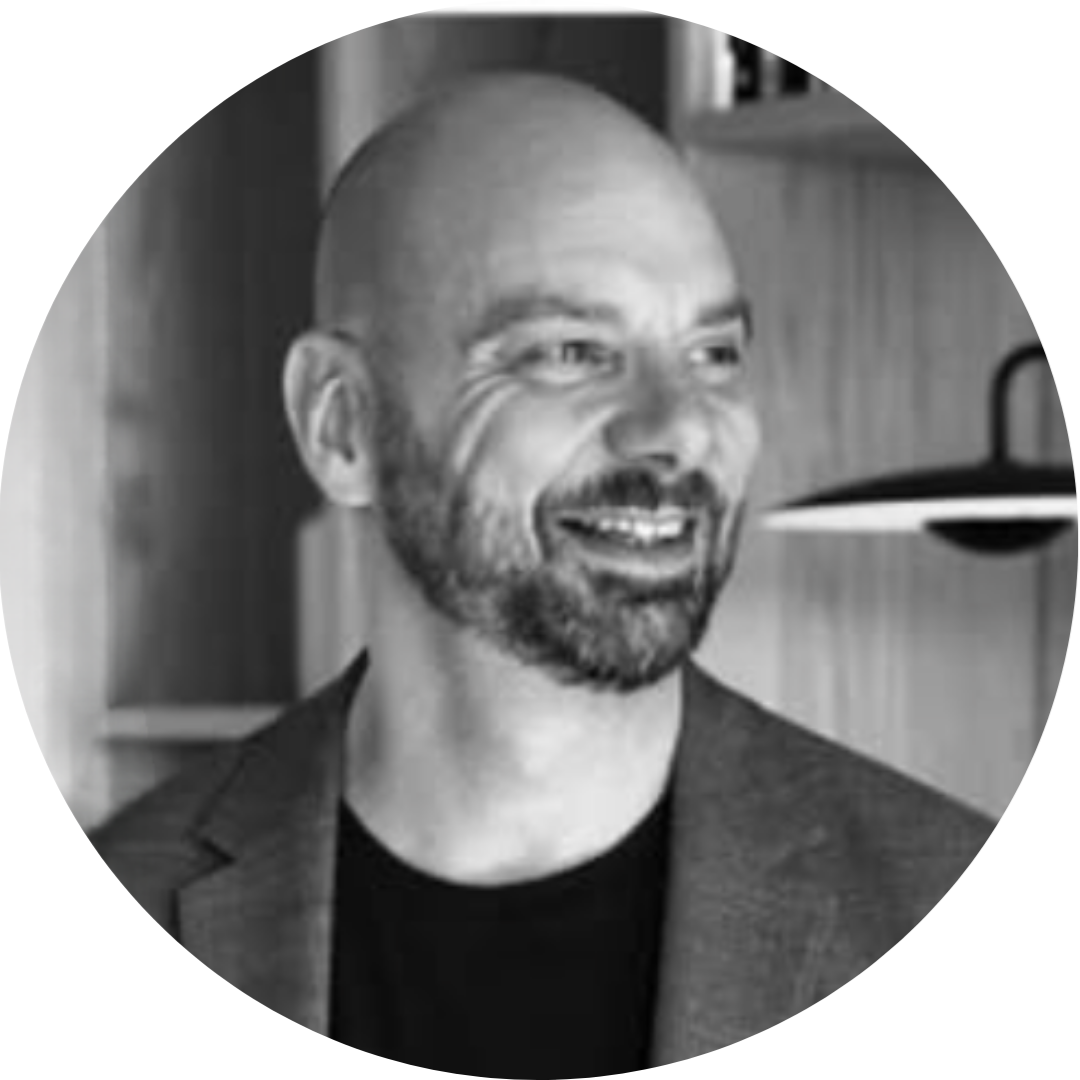 A black-and-white photo of a smiling, bald man with a beard, wearing a blazer, in an indoor setting.