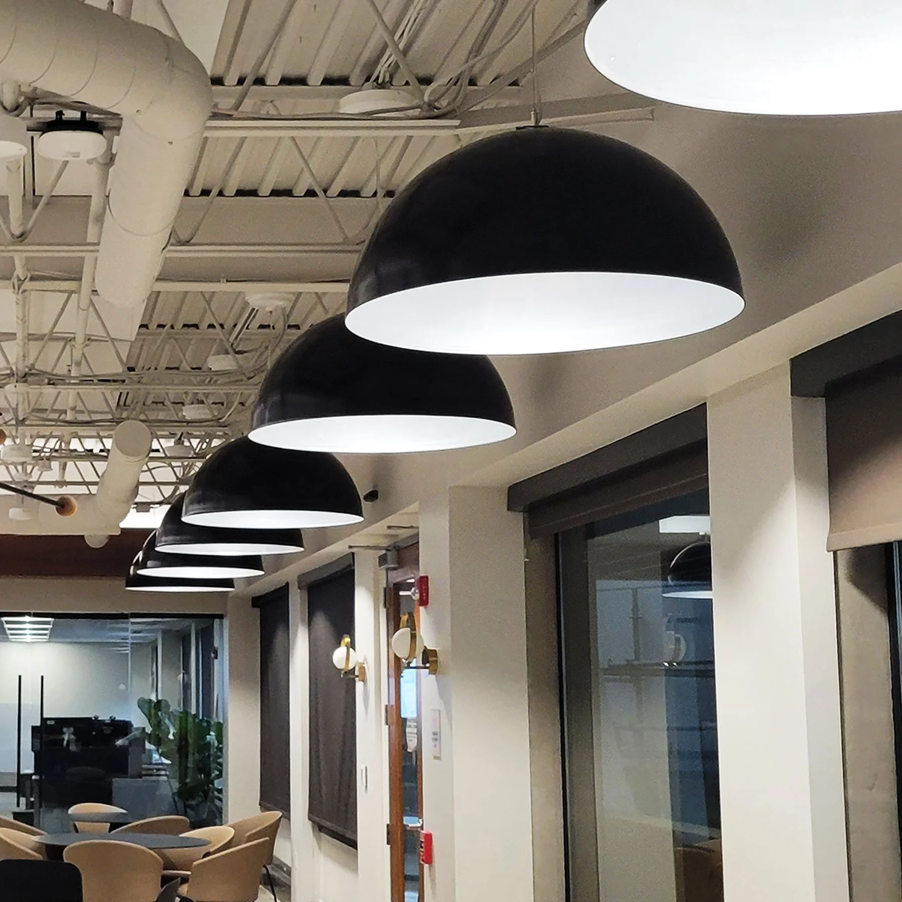 Row of oversized black dome pendant lights with white interiors hanging above an open-plan office corridor