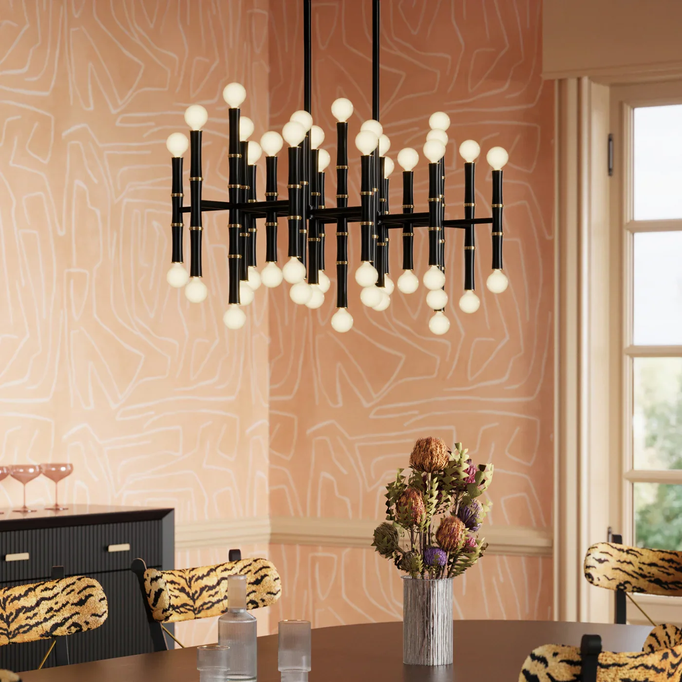 A modern chandelier with multiple small white spherical lights hangs above a round dining table in a room with peach-colored walls with geometric white line patterns. A vase with a bouquet of dried flowers is on the table, and chairs with tiger-stripe patterned upholstery surround it.