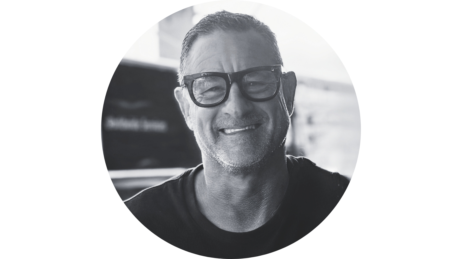 A smiling man with glasses in a black and white photo, wearing a dark shirt, in an indoor setting with windows in the background.