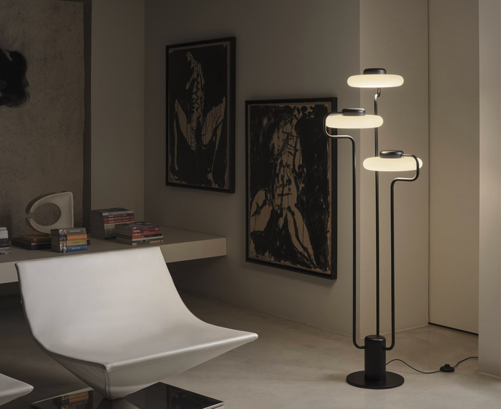 A modern living room with a white curved lounge chair, a white table with stacked books, abstract black and beige artwork on the wall, and a black floor lamp with three illuminated oval-shaped lights.
