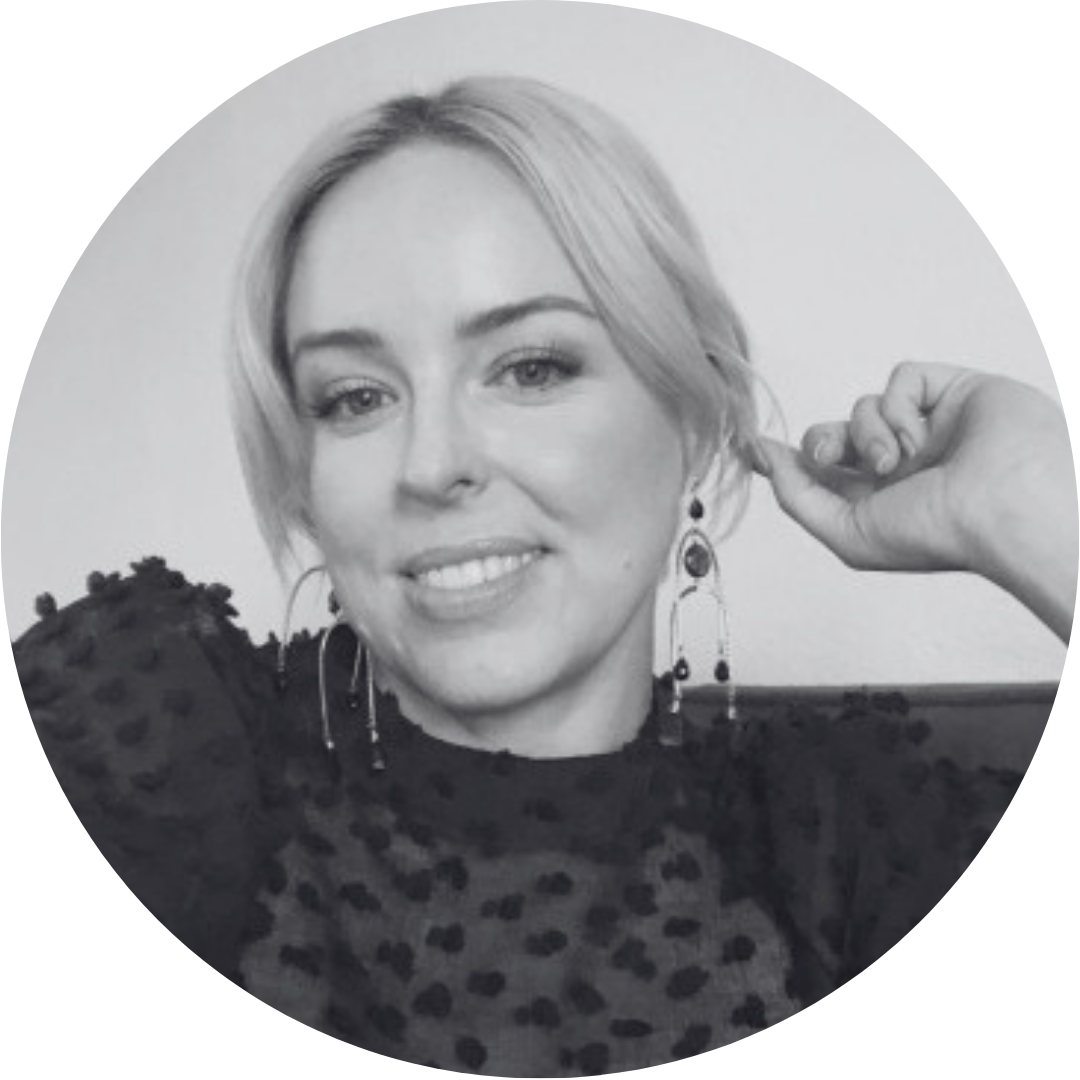 A woman with short blonde hair smiling, wearing earrings, in a leopard print top, touching her ear, in a grayscale photo.
