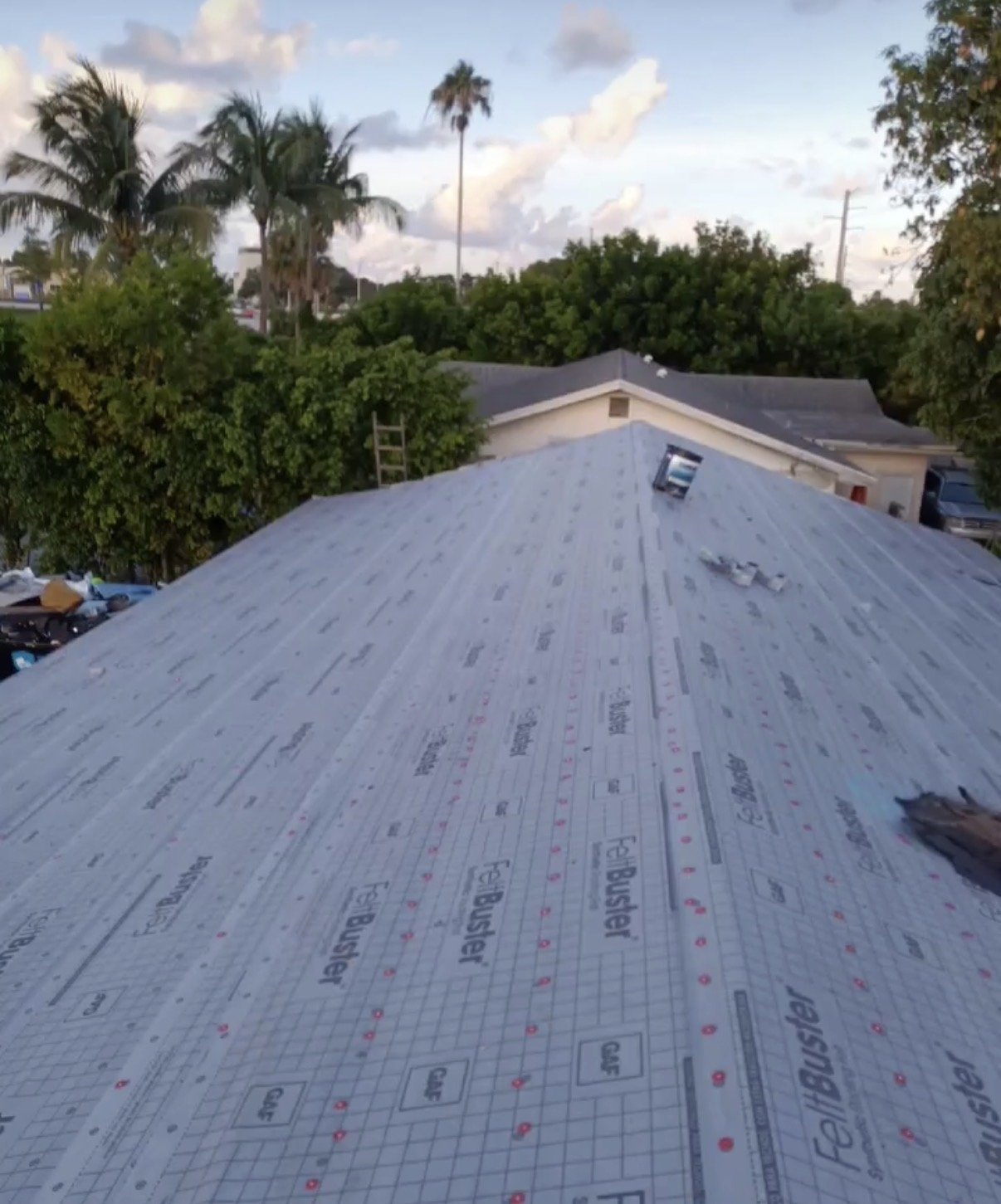 View of a roof under construction with weatherproofing underlayment, surrounded by trees and houses, and a sky with clouds in the background.