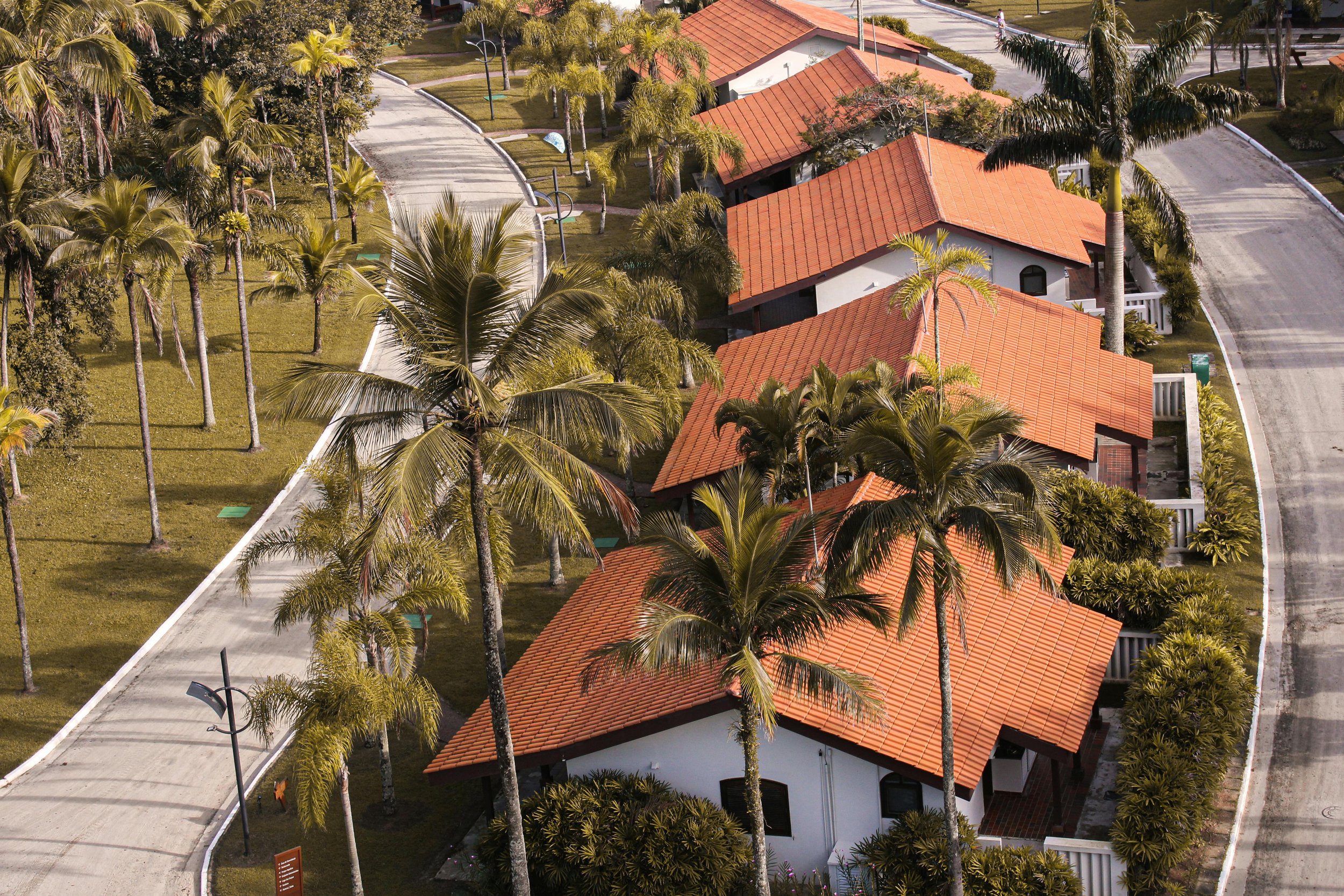 Aerial view of a neighborhood with white houses that have red-tiled roofs, surrounded by green trees and palm trees, with a curved sidewalk and street.
