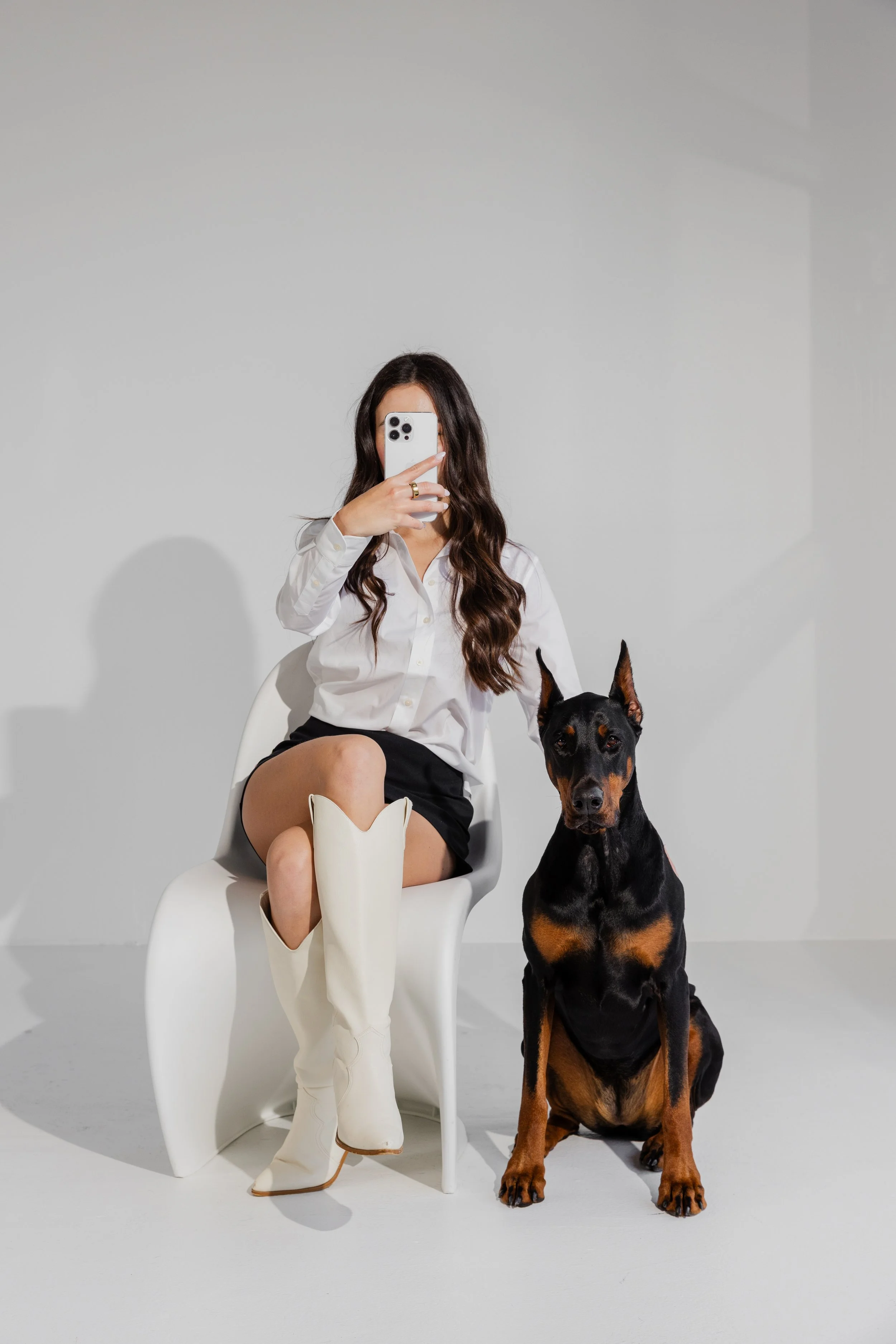 A woman with long wavy brown hair wearing a white shirt and black skirt, sitting on a white chair, taking a selfie with her smartphone. A Doberman dog is sitting next to her.