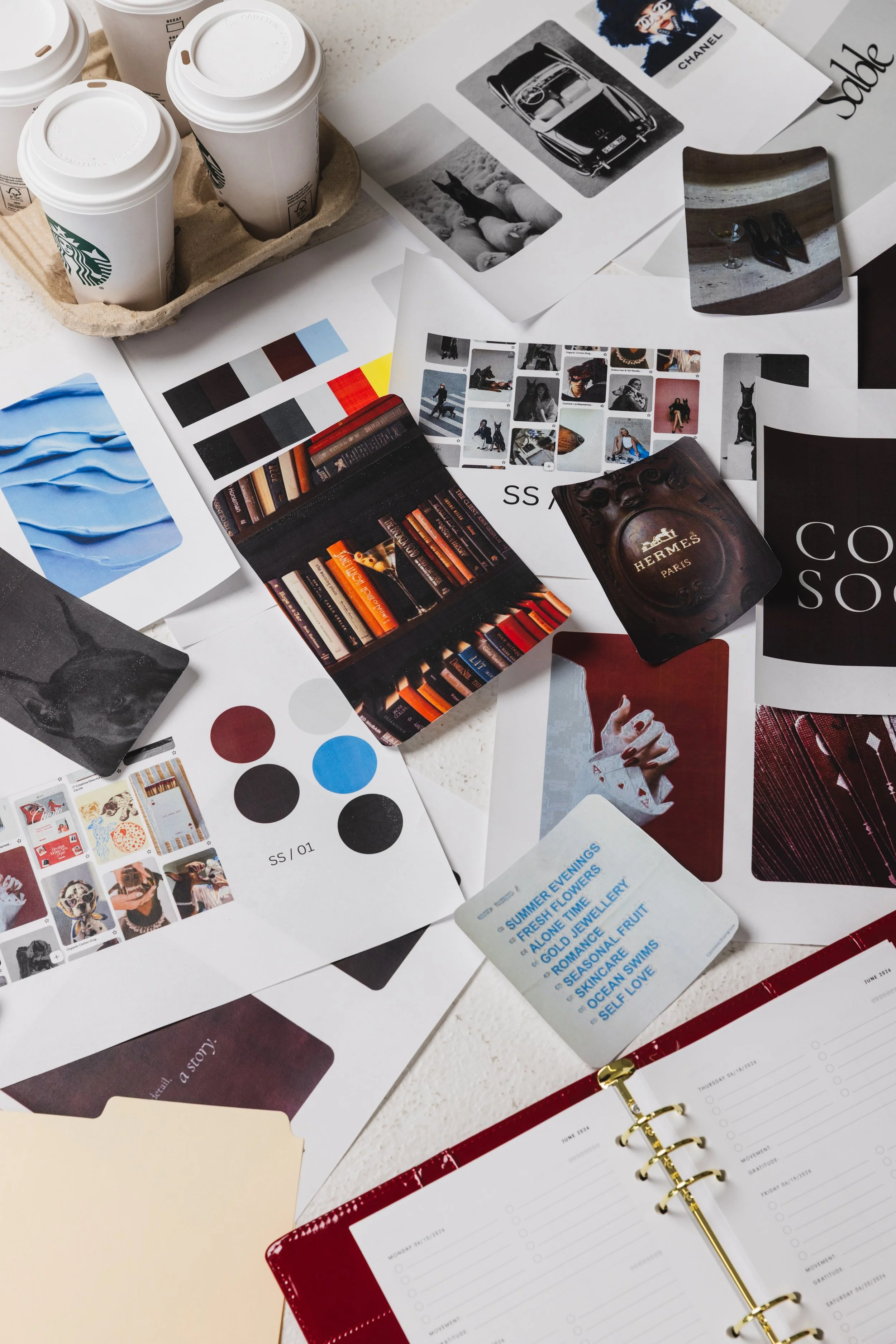A cluttered desk with disposable coffee cups, printed images, color swatches, bookshelves, playing cards, and a red planner with a gold pen.