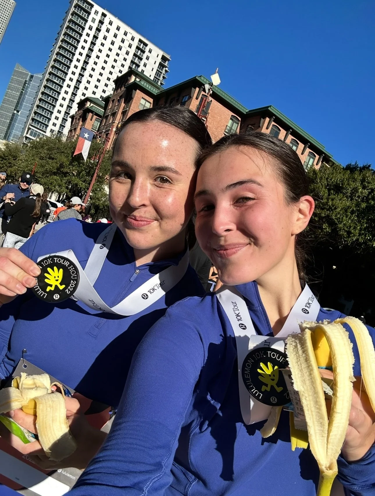 Two young women wearing blue athletic gear with medals around their necks, holding peeled bananas, smiling outdoors after a race in an urban setting with buildings and blue sky in the background.