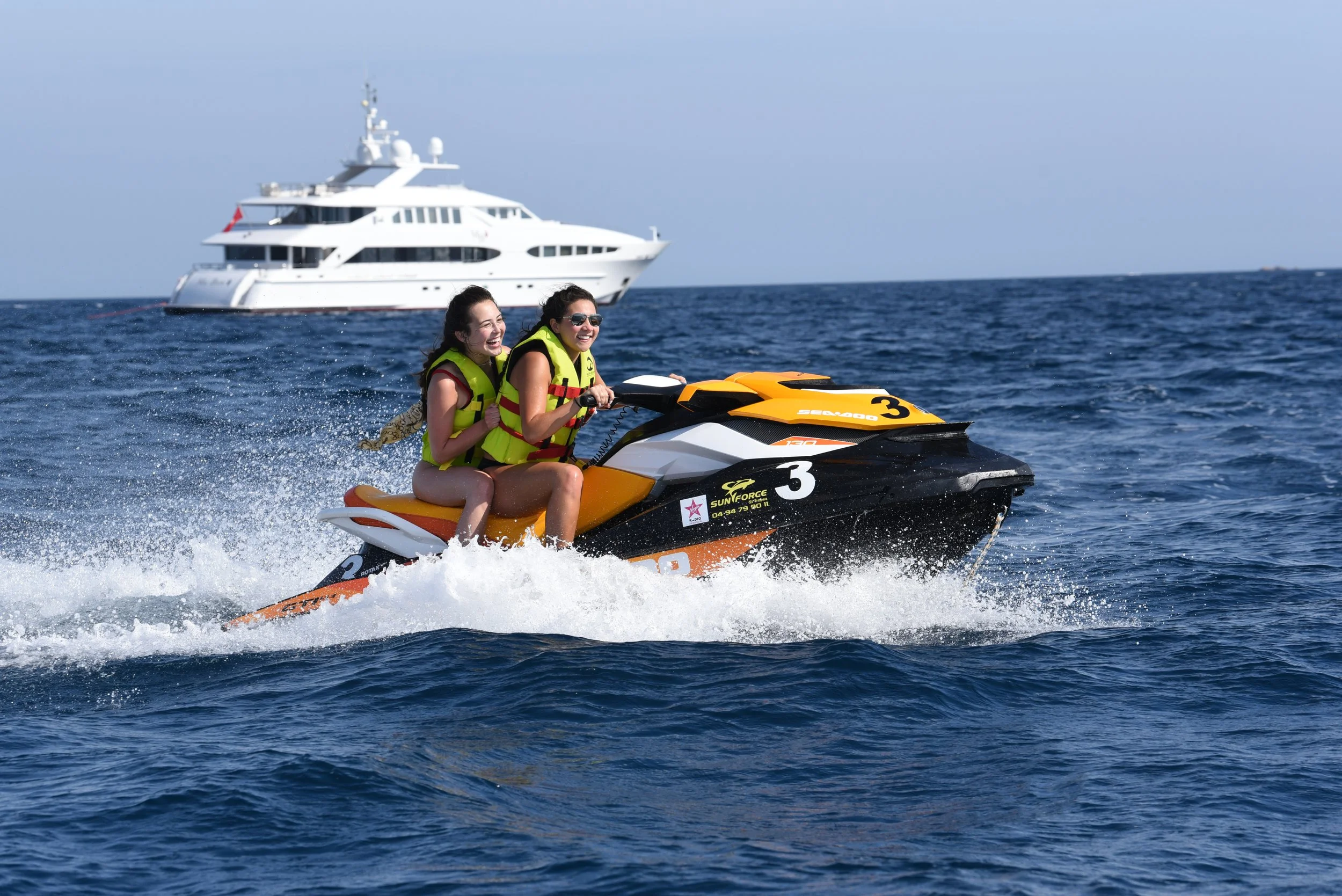 Two women wearing life jackets riding a yellow and black jet ski on the water while a large white yacht is in the background.