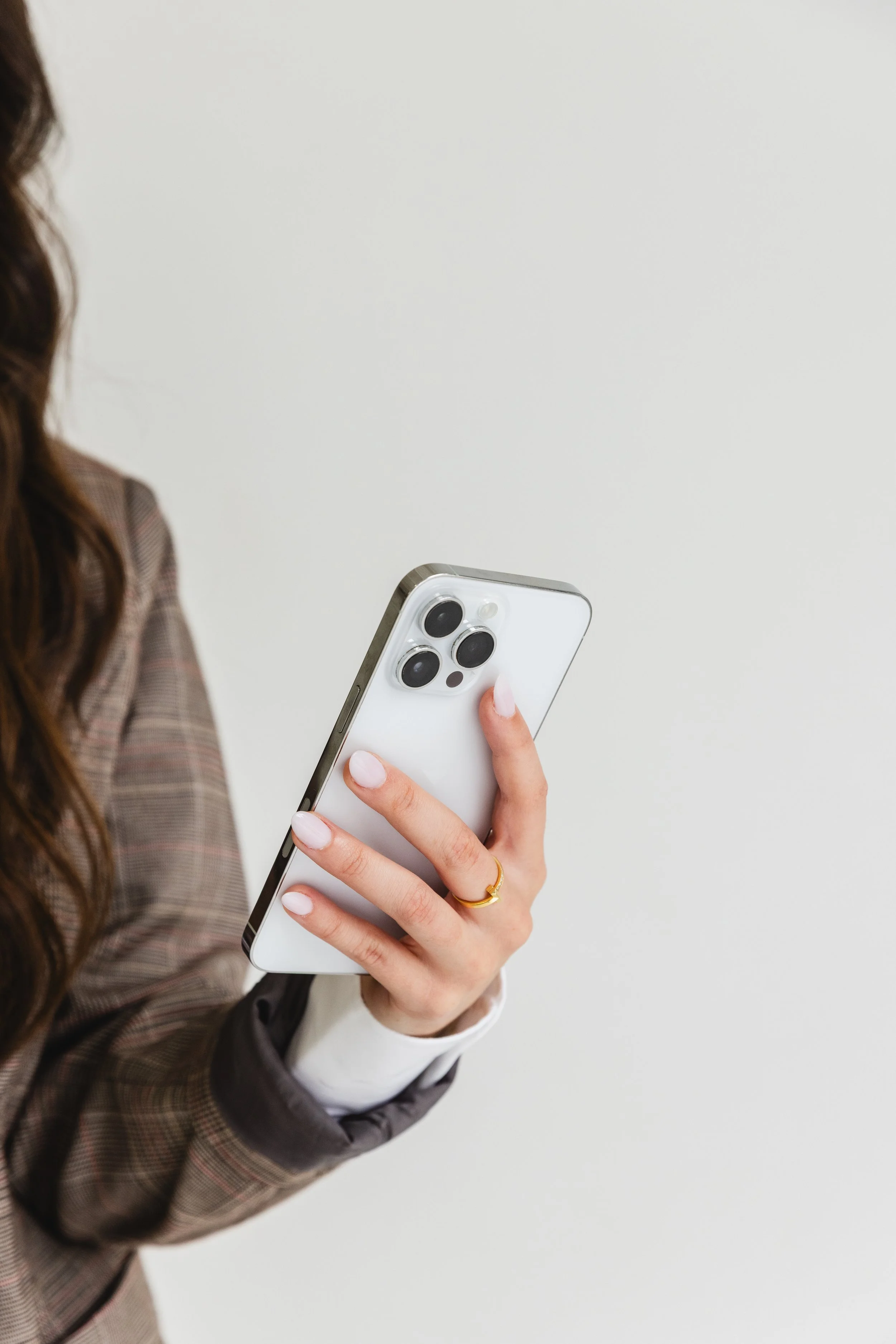 A person holding a smartphone with a triple-camera setup, dressed in a plaid blazer with a white shirt underneath, standing against a plain white background.
