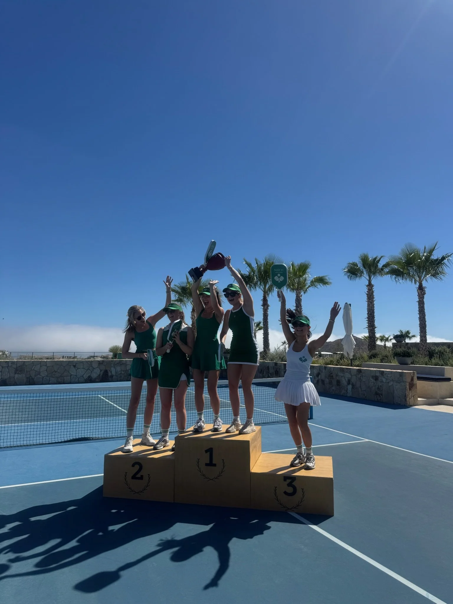 Women standing on a winners' podium with a background of palm trees and a tennis court, celebrating their victory, under a clear blue sky.