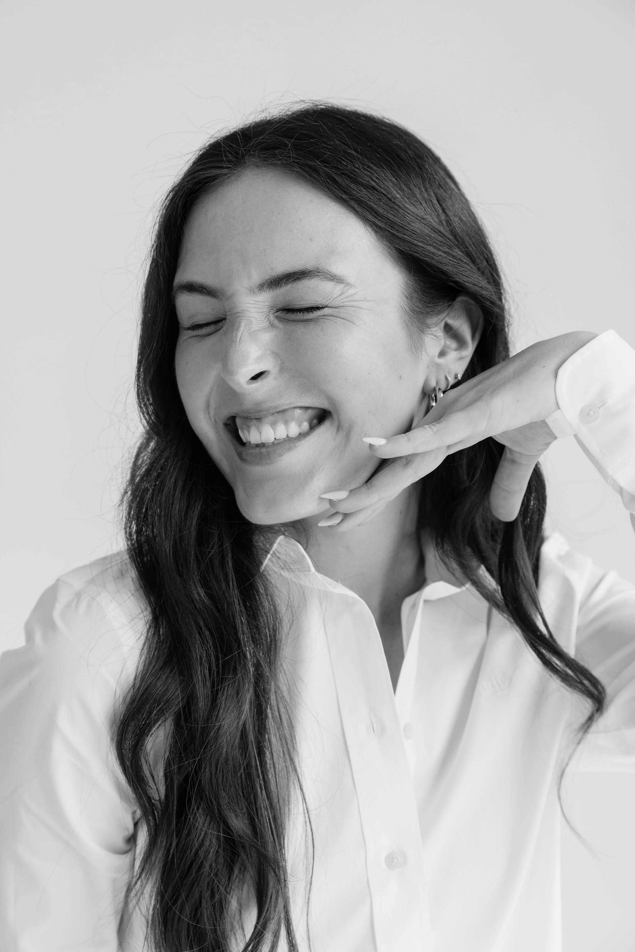 A woman with long dark hair smiling with her eyes closed, wearing a white shirt, touching her chin with her hand.