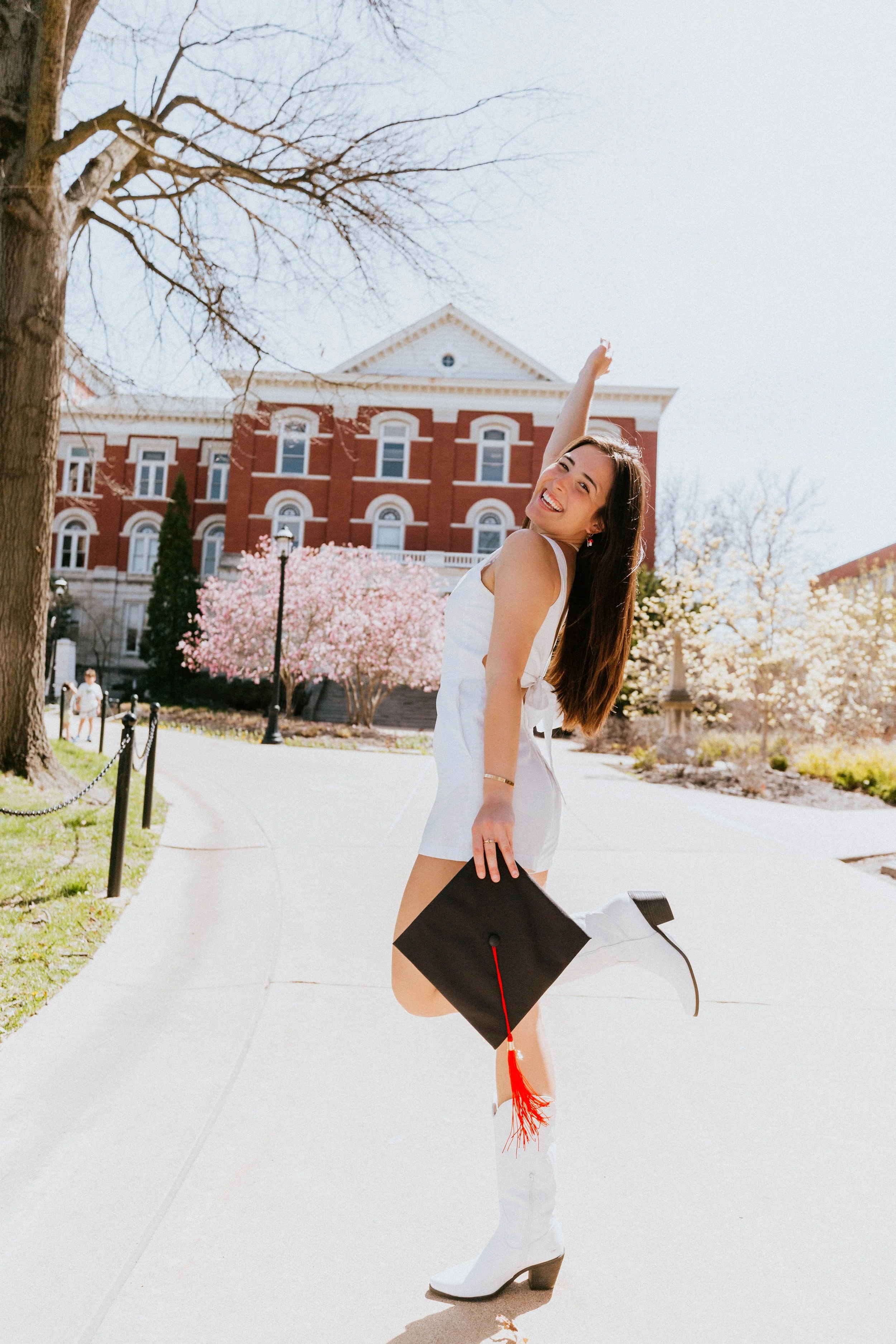 A young woman in white boots and a white dress holding a black graduation cap with a red tassel, smiling and raising her arm, standing on a sidewalk with blossoming trees and a large historic brick building in the background.