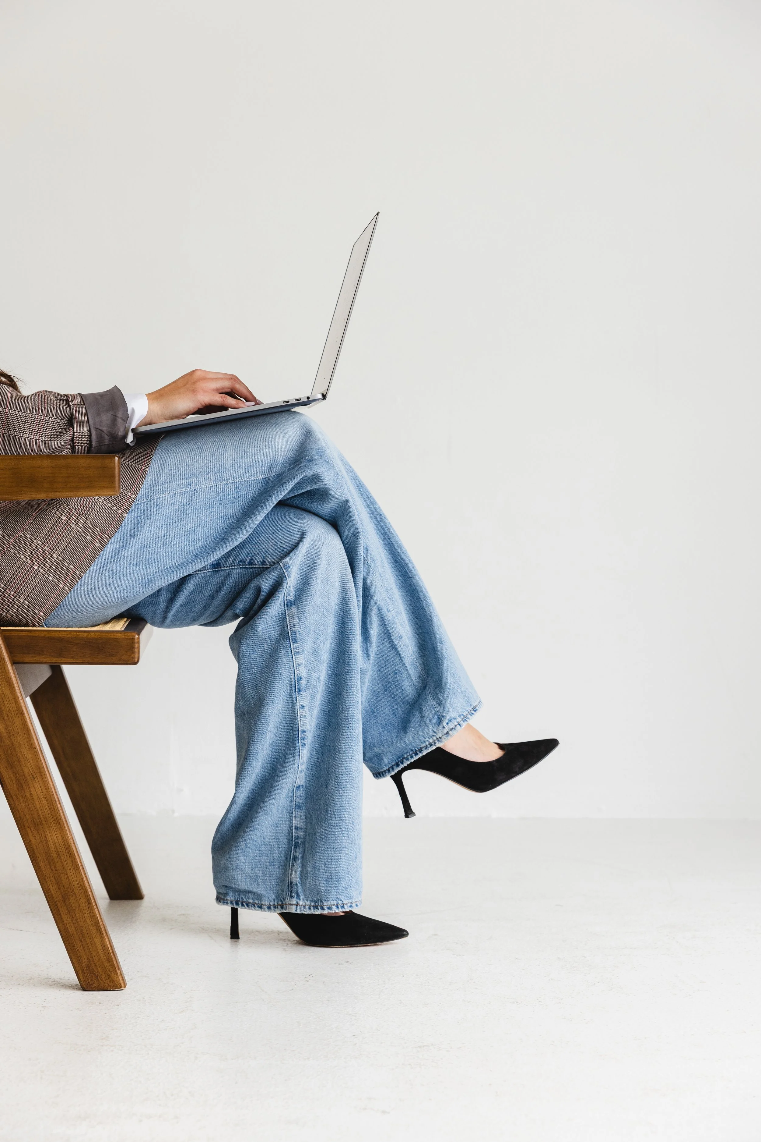 A woman sitting on a wooden chair, wearing black high heels, light blue jeans, a checkered blazer, and a white shirt, with her left leg crossed over her right, typing on a silver laptop against a plain white wall background.