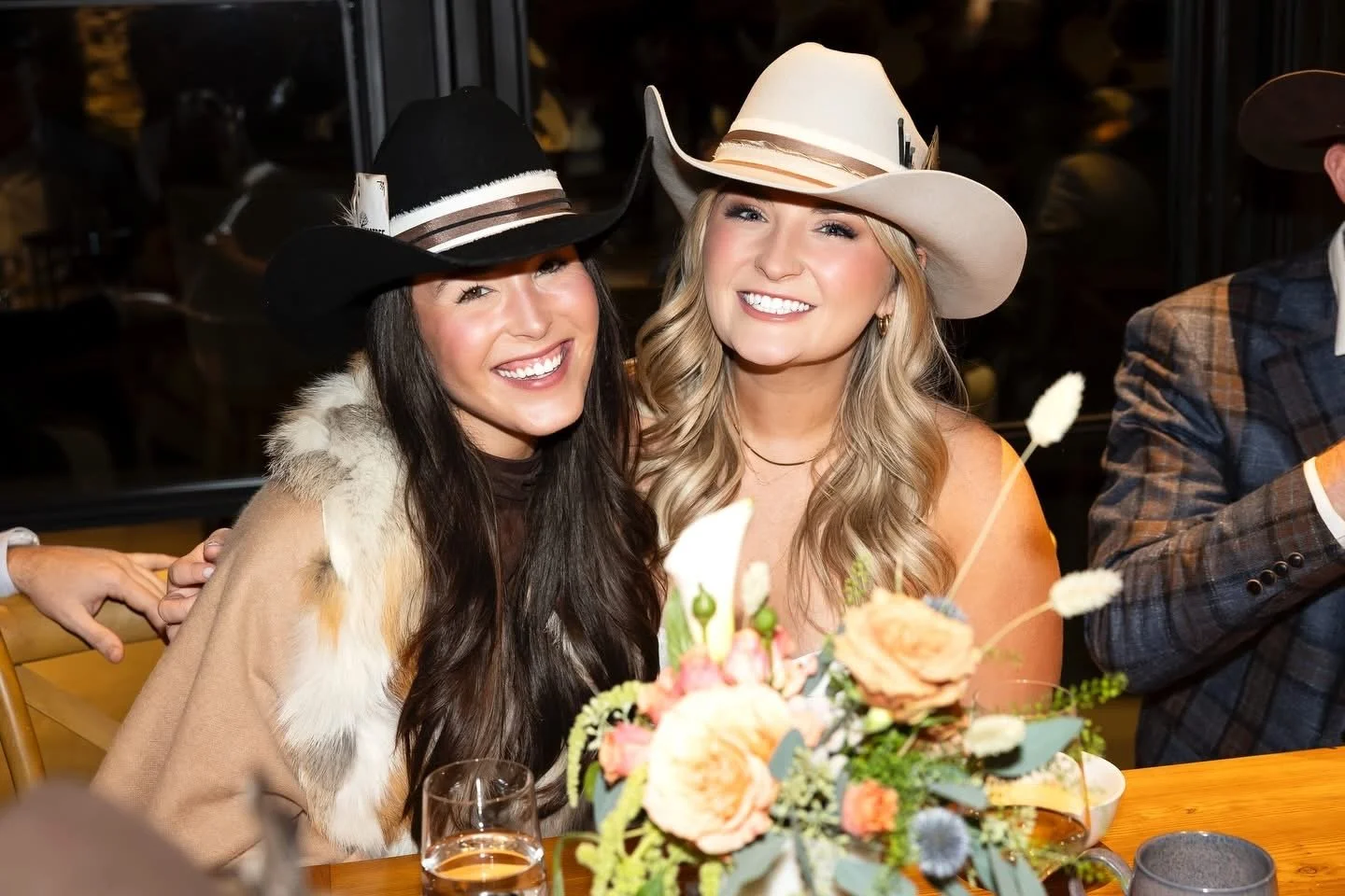 Two women wearing cowboy hats smiling at a table with a bouquet of flowers, a glass of drink, and a dark background.