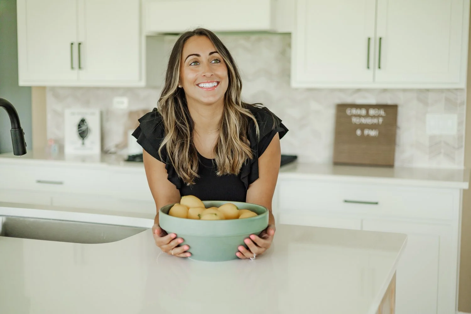 A woman with long, wavy hair and a black top, smiling, holding a large green bowl of peaches in a modern kitchen.