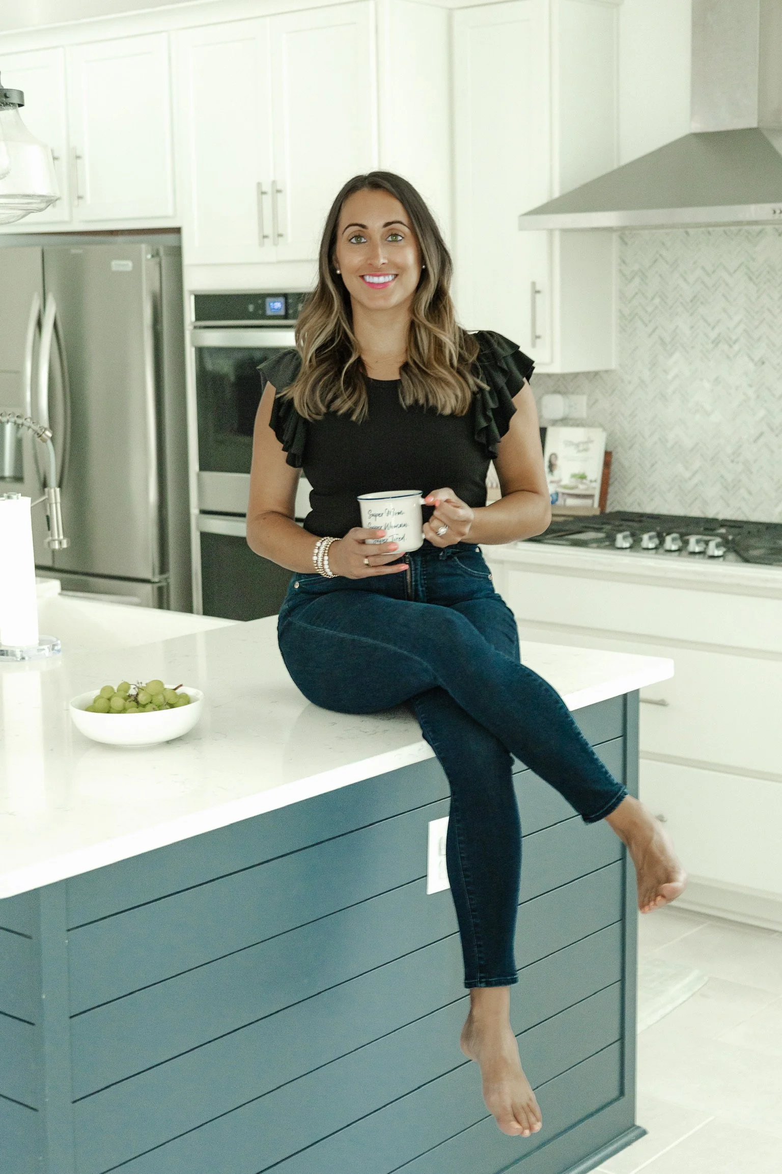 A woman with long brown hair in a black ruffled sleeve top and blue jeans is sitting on a kitchen island with one leg crossed over the other and her bare foot hanging down. She is holding a white mug and smiling at the camera. The kitchen has white cabinets, a stainless steel refrigerator, and a black oven. There is a bowl of green grapes on the island.