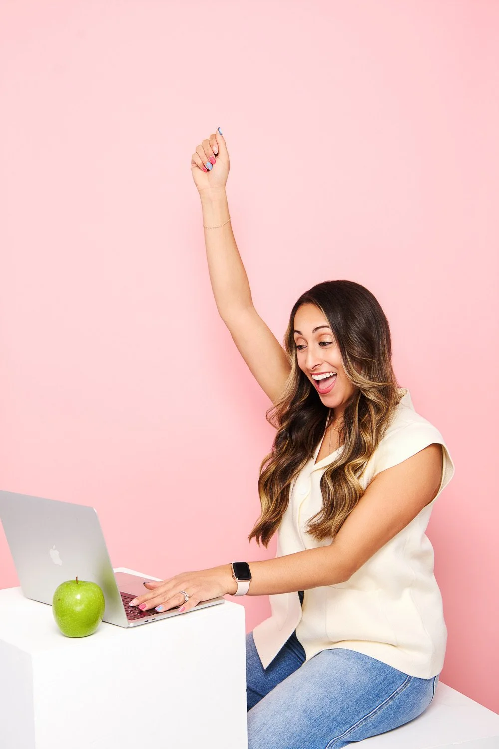 A woman celebrating while working on a laptop with a green apple on a white table against a pink background.