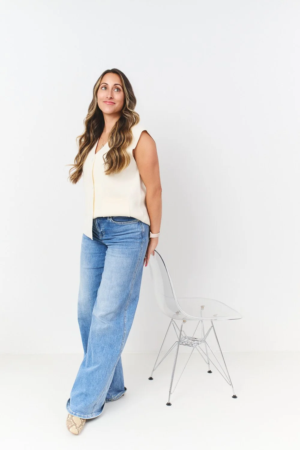 A woman with long wavy hair wearing a sleeveless cream top and blue jeans standing next to a clear modern chair in a white room.