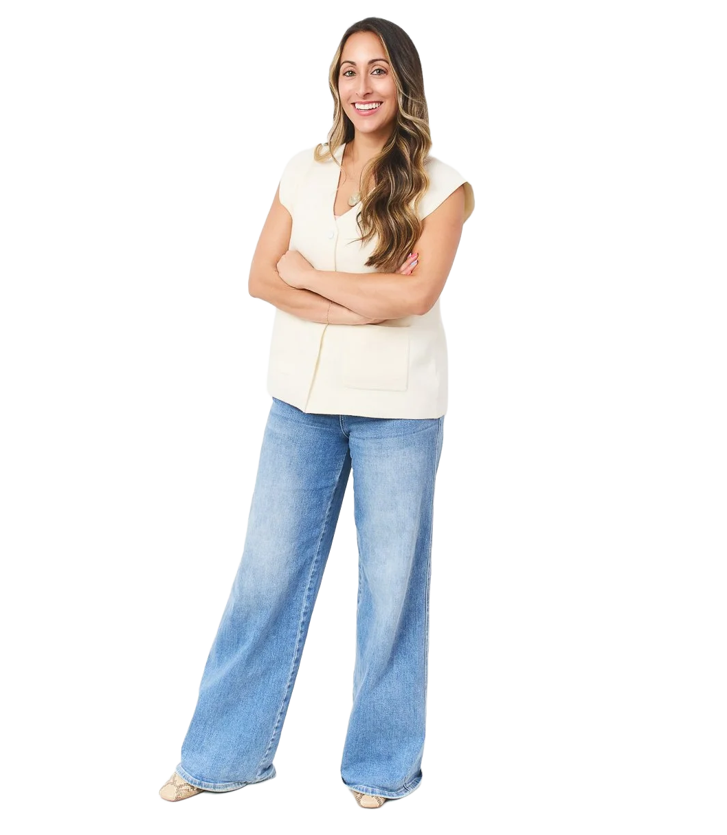 A woman with long wavy hair smiling and standing with arms crossed, wearing a cream-colored top and wide-leg jeans against a plain white background.