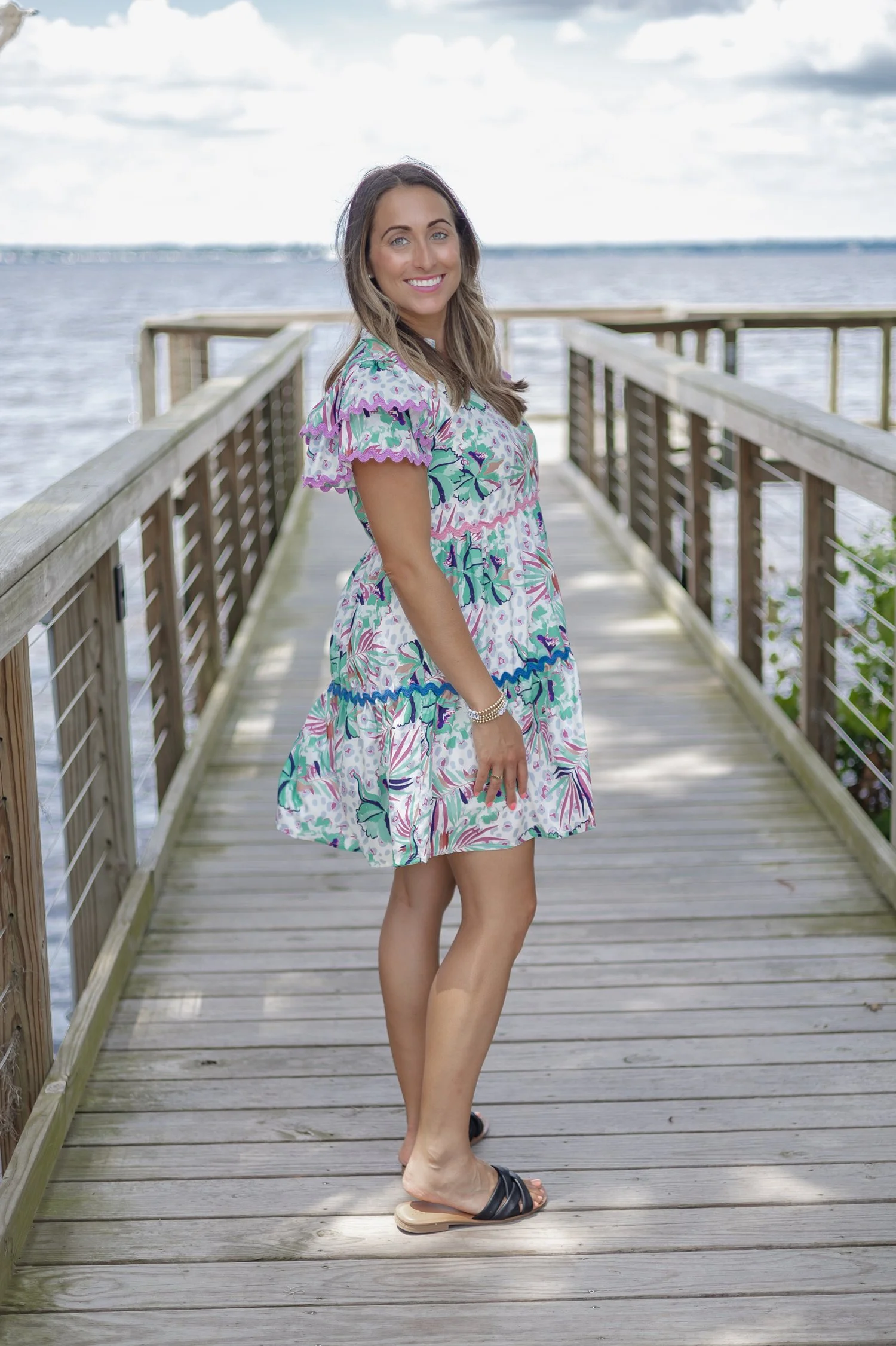 A woman standing on a wooden pier with water in the background. She is smiling and wearing a colorful floral dress and black sandals.