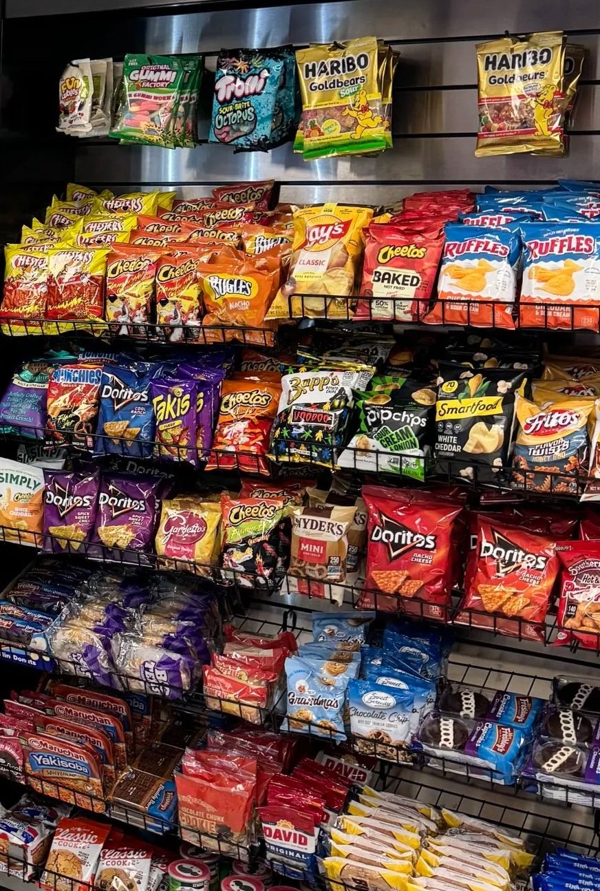 Shelves of various snack bags in a store, including potato chips, cheese puffs, pretzels, and candy, organized in metal baskets and on hooks.