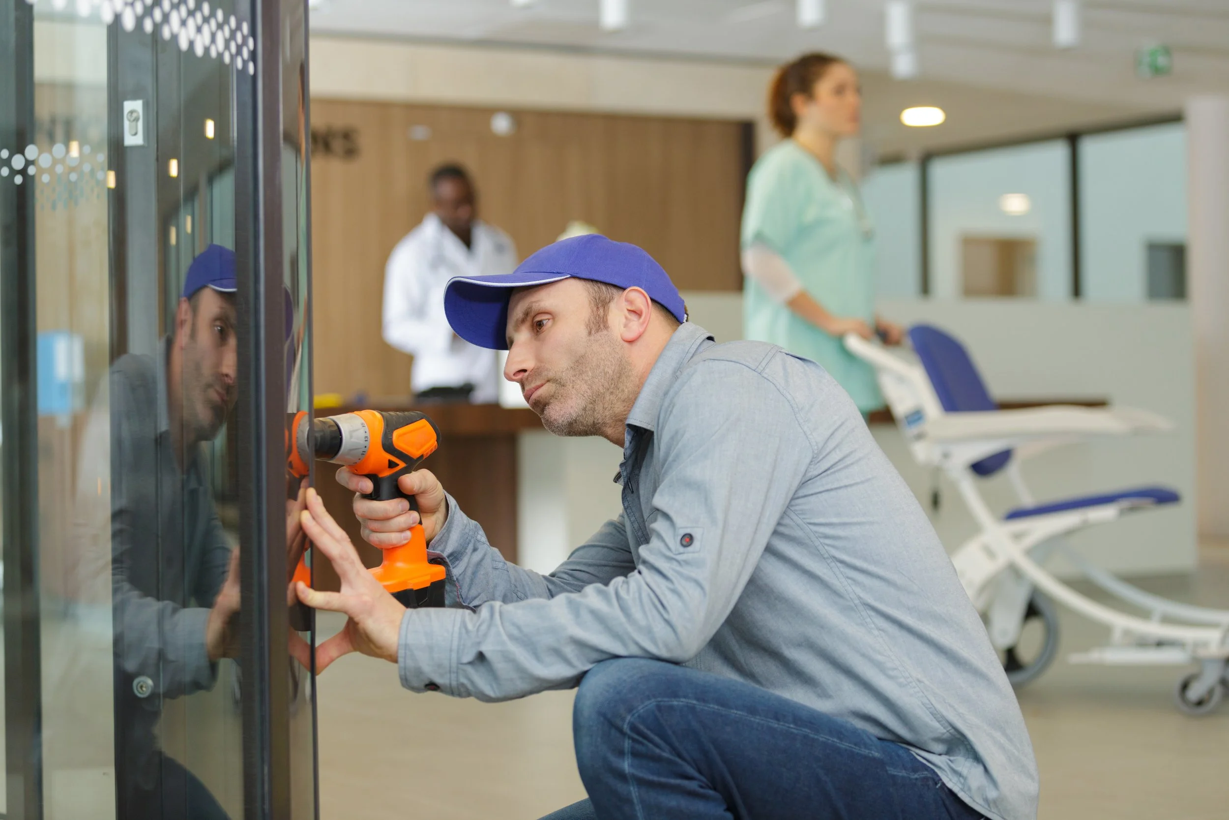 Man wearing a blue cap repairs a glass window with a tool in a medical or healthcare facility, with medical staff and beds visible in the background.