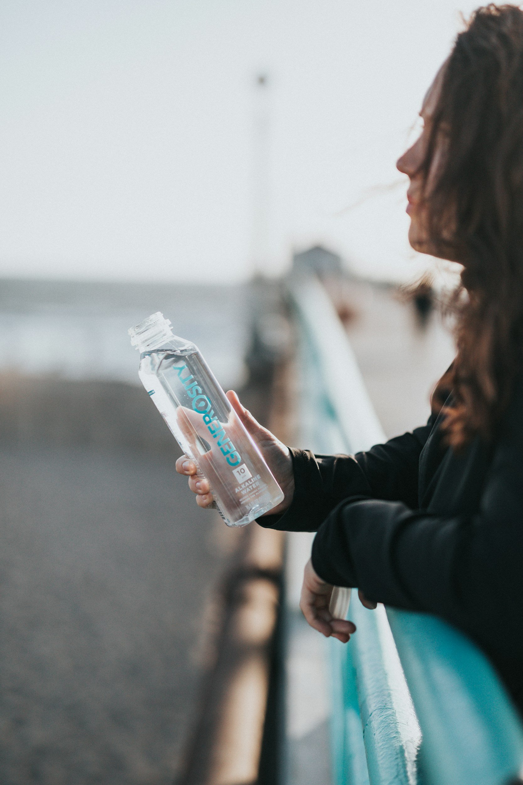 A woman in a black jacket holding a clear plastic bottle of carbonated alkaline water while leaning on a blue railing at the beach.
