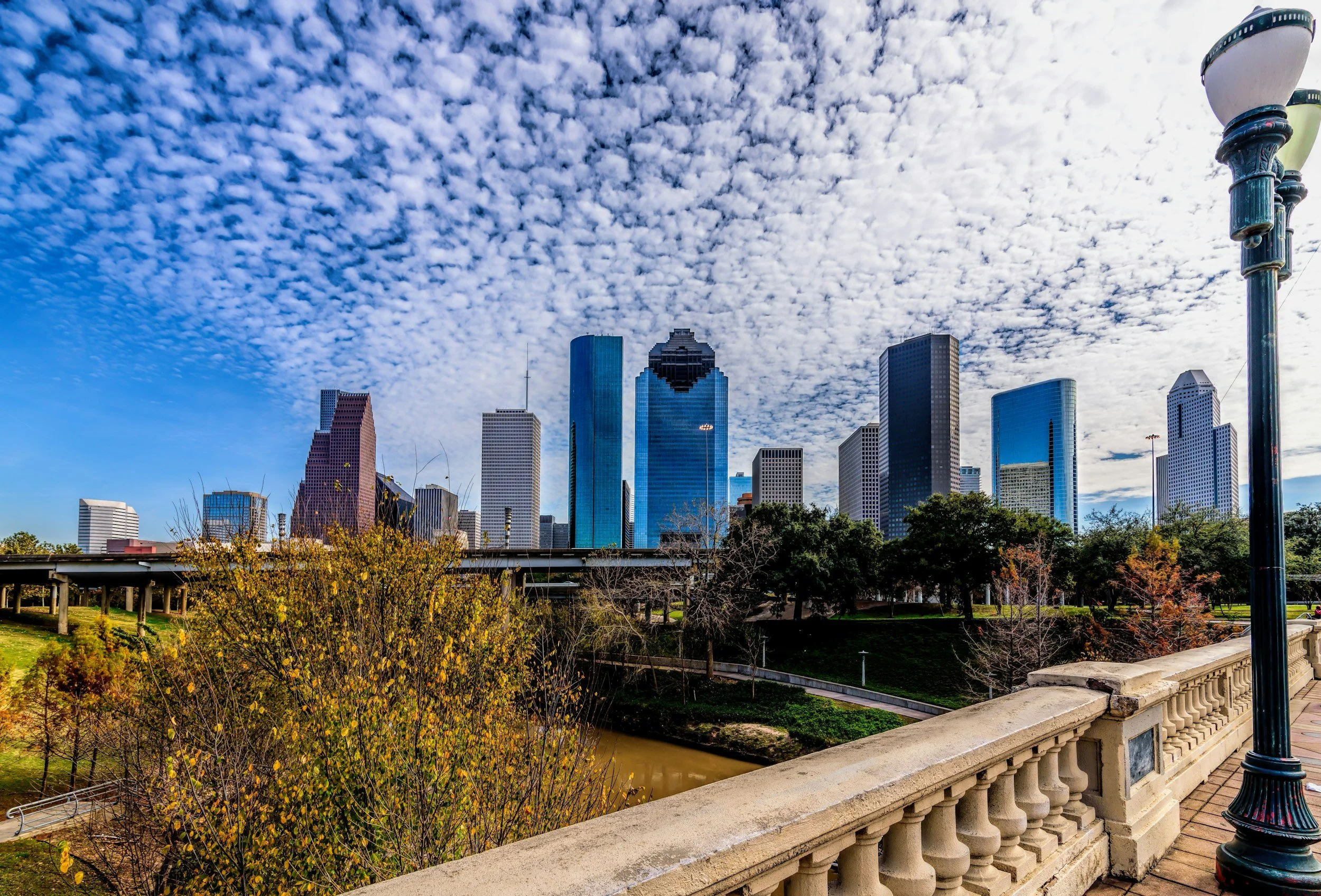 City skyline with tall modern buildings under a cloudy sky, viewed from a park with a stone railing and decorative lamp post in the foreground.