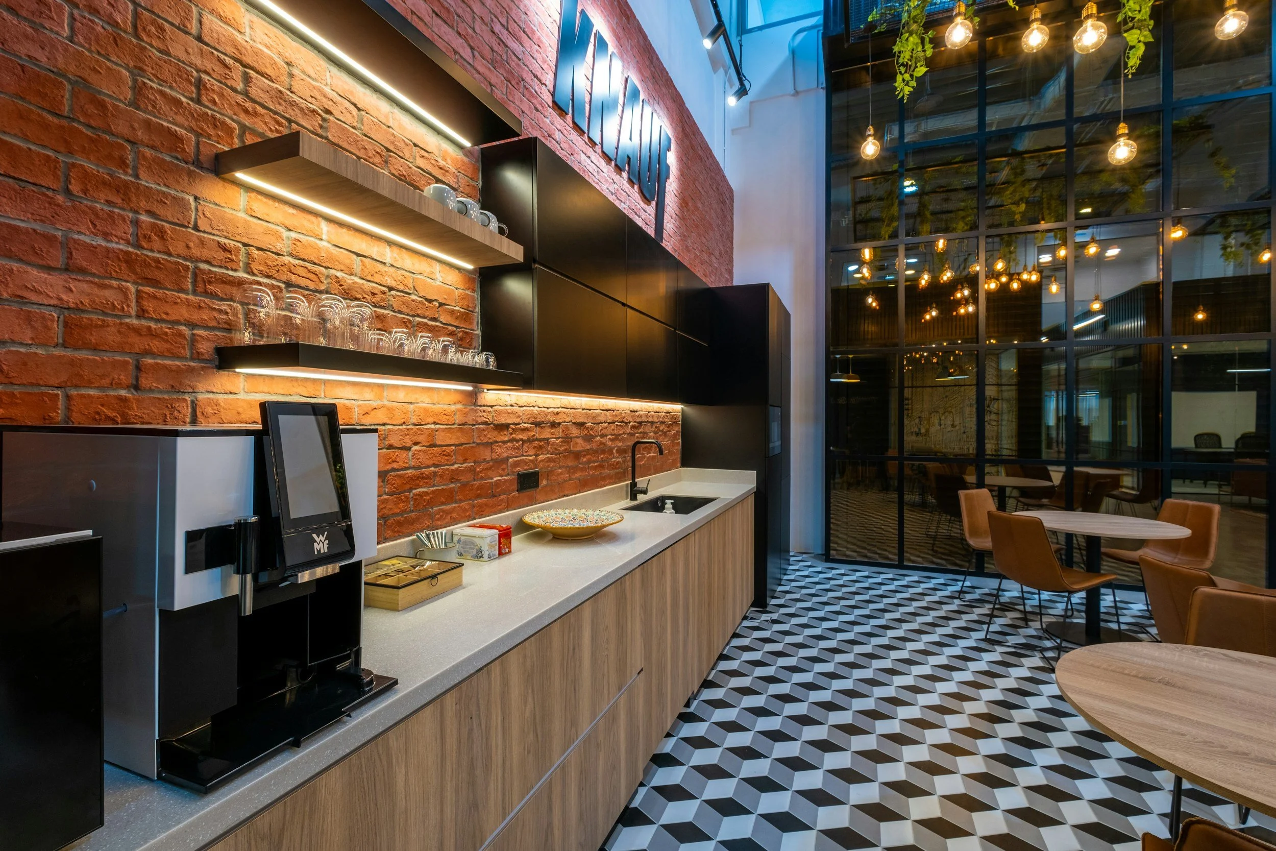 Modern café interior featuring a brick accent wall with shelves, black cabinetry, a white countertop with cups and a coffee machine, and large windows with warm hanging string lights and green plants outside. Seating includes chairs and tables with geometric floor tiles.
