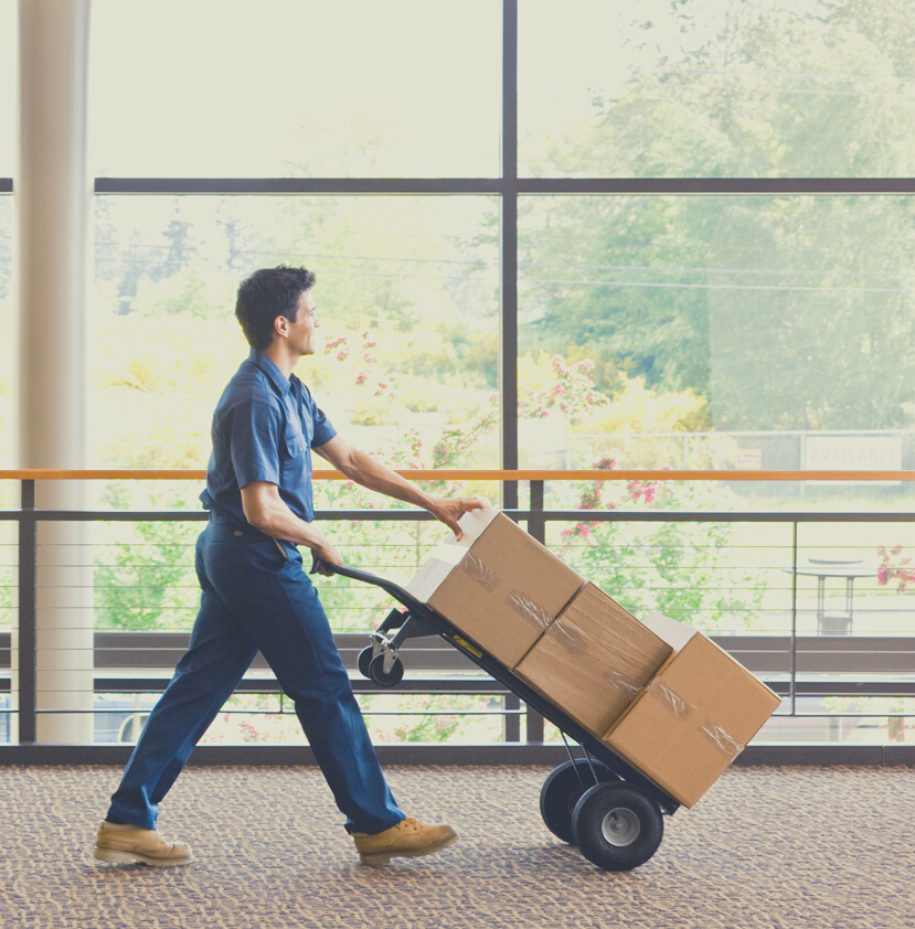 A person wearing blue scrubs pushing a cart with boxes inside an indoor space with large window and view of trees outside.
