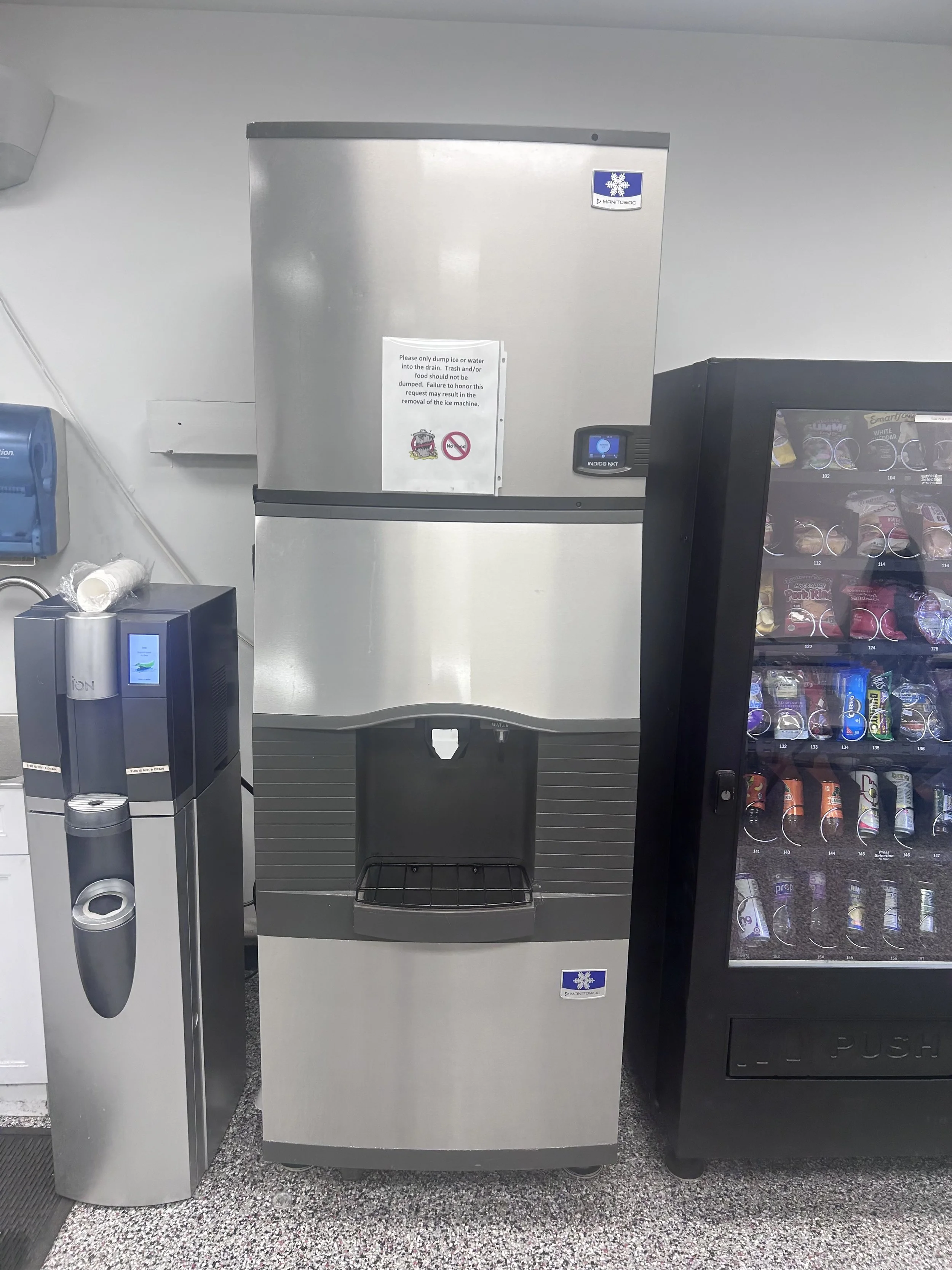 A stainless steel ice machine with a digital control panel, surrounded by a vending machine with snacks to the right, a water dispenser to the left, and a silver paper towel dispenser on the wall, in a retail or break room setting.
