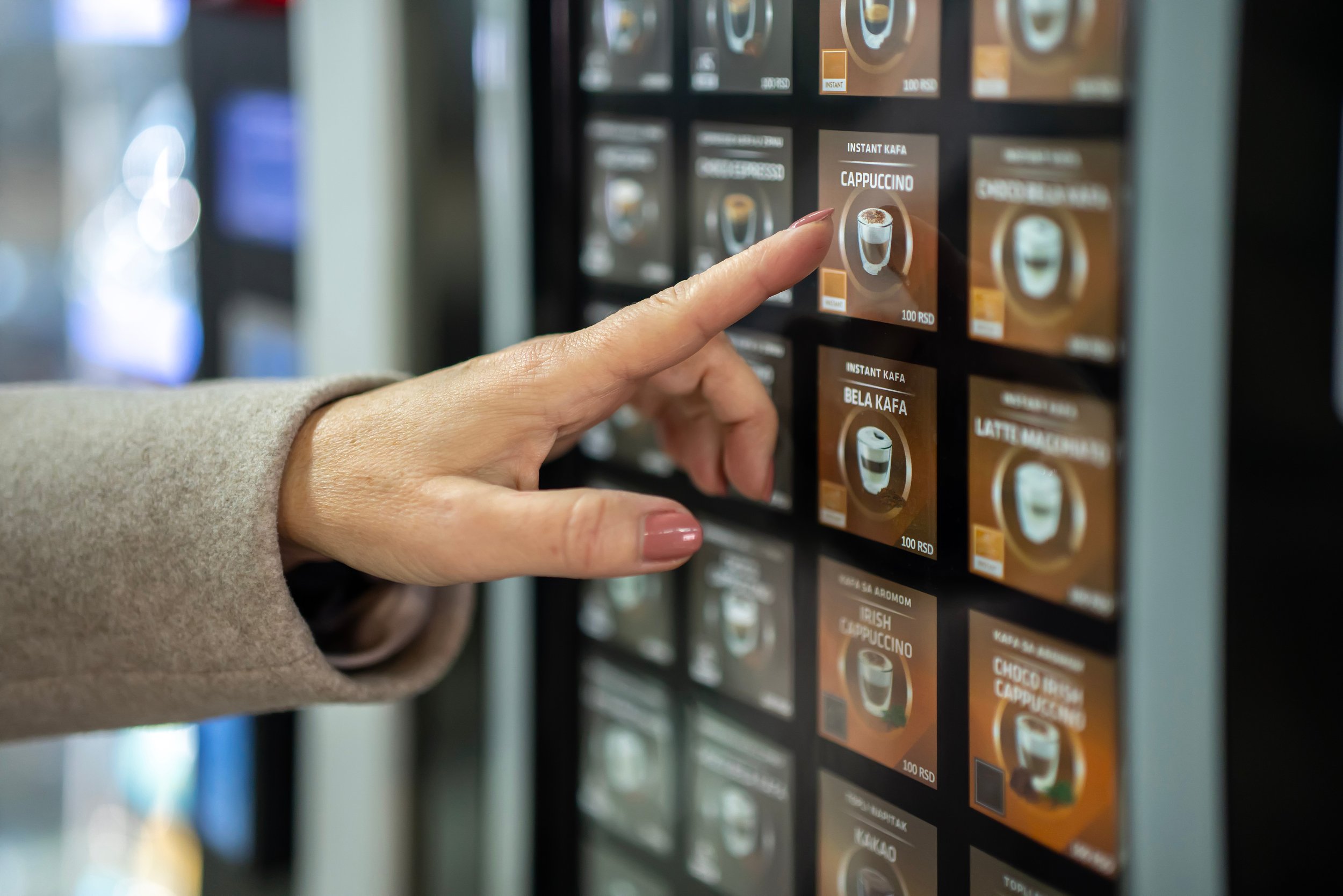 Close-up of a person's hand selecting a coffee flavor from a vending machine. The flavor is labeled 'Cappuccino' and has an image of a coffee cup on the button.