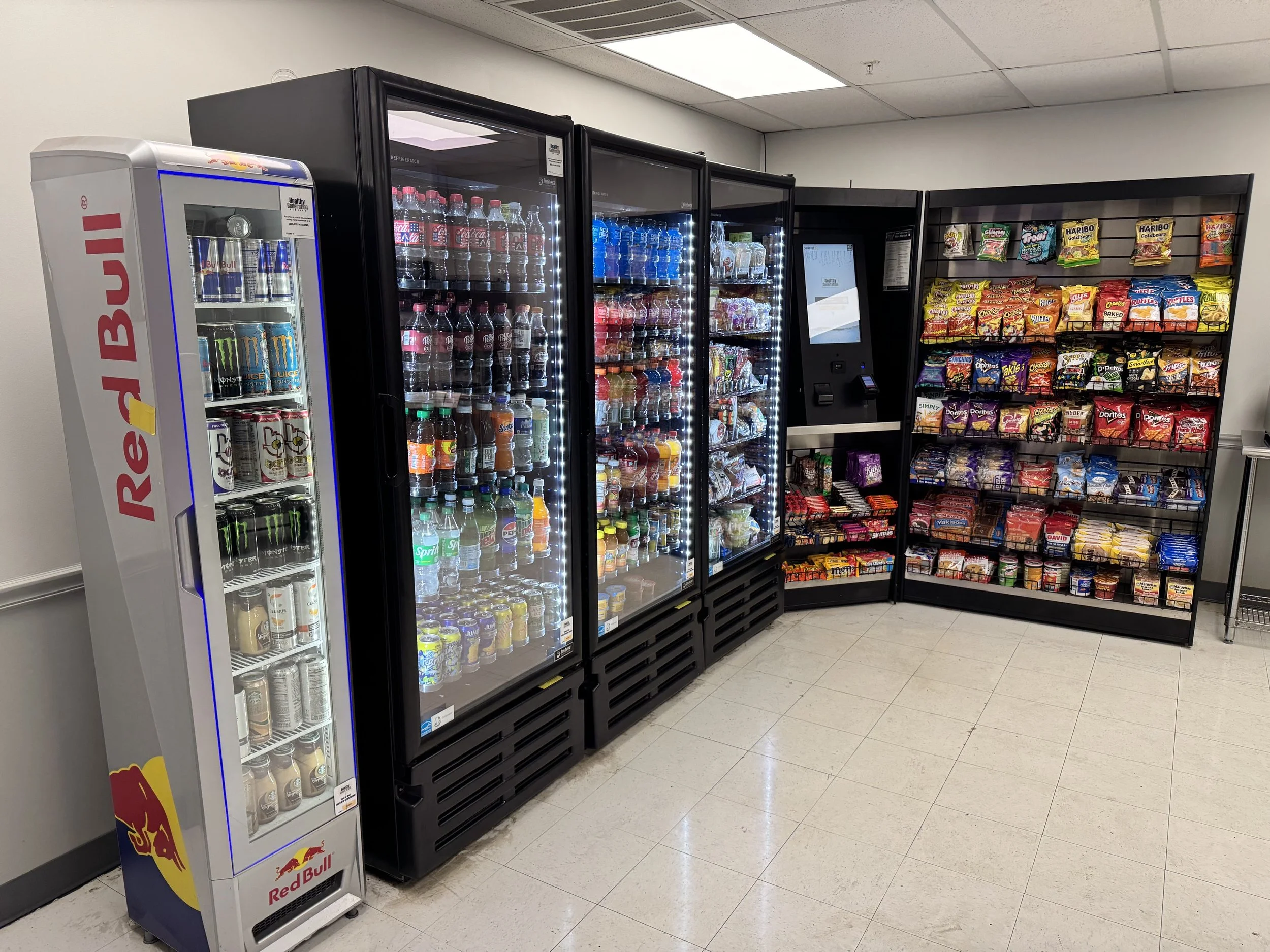 A vending machine with Red Bull energy drinks, several glass door refrigerators filled with bottled beverages, and a snack display with various chips and candy in a convenience store.