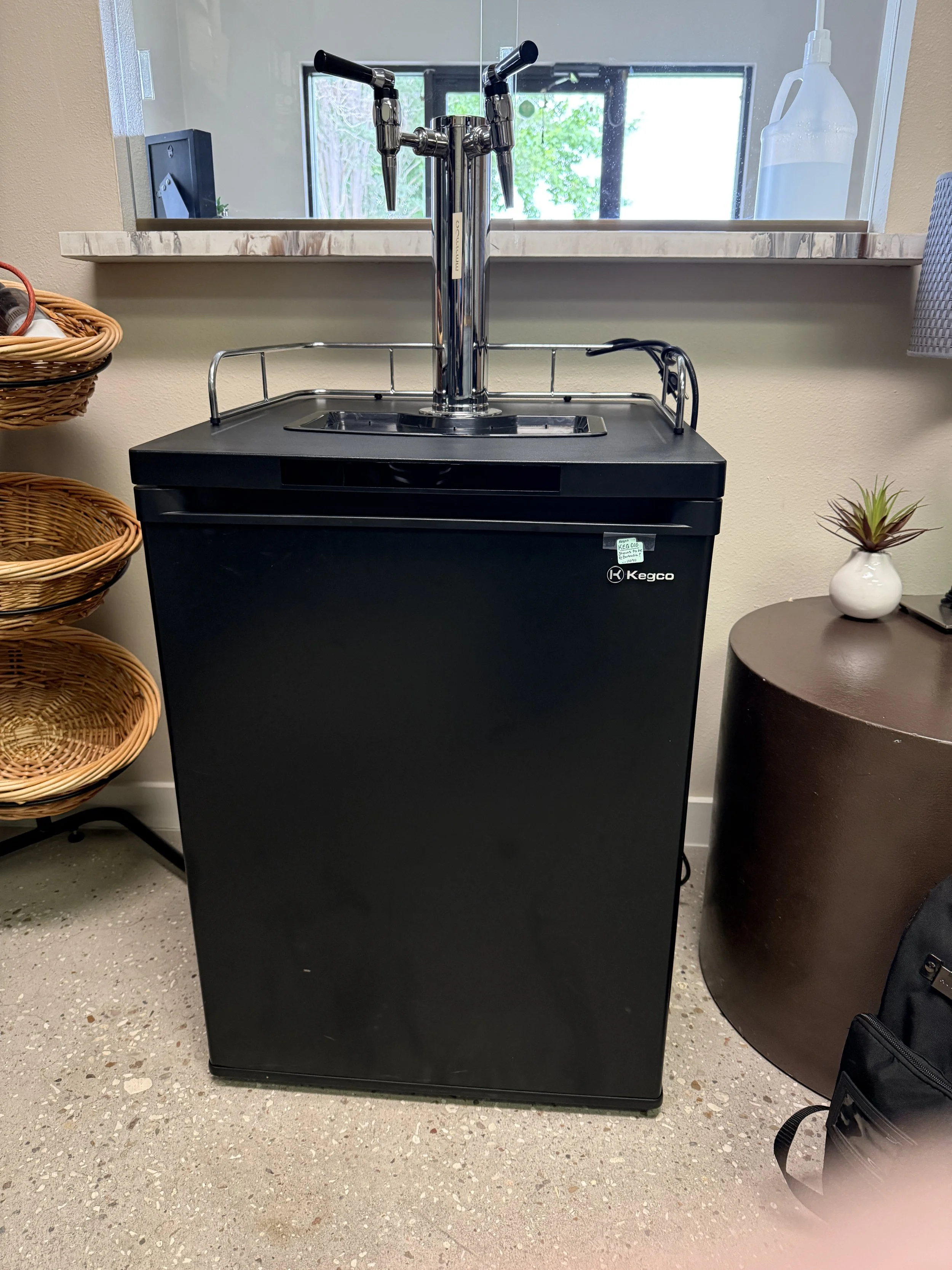 A black Kegco beer dispenser with dual taps, positioned next to a small round table with a potted plant, and a white vessel in the background behind a window.