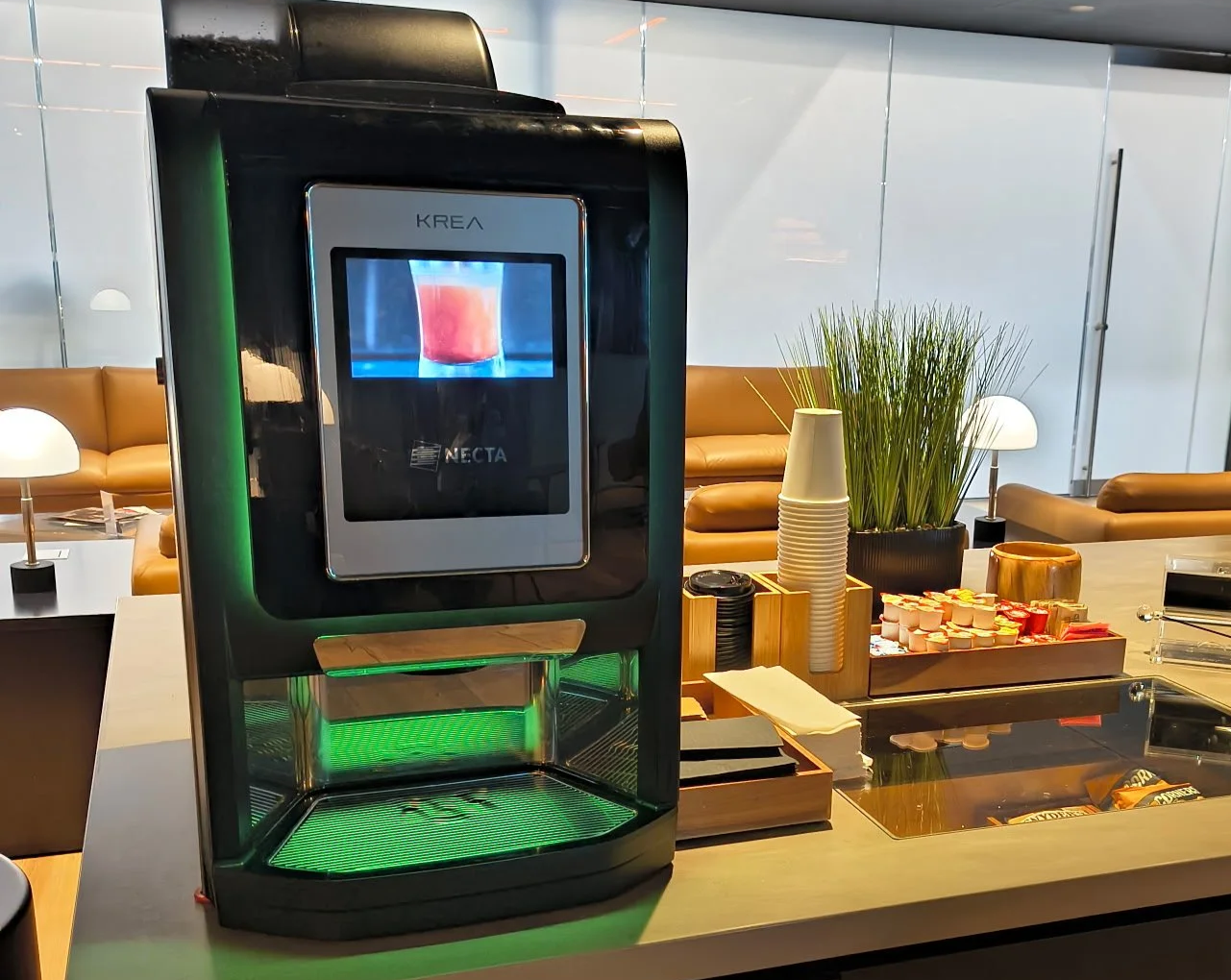 A beverage dispensing machine on a counter in a lounge or office setting, with cups and snacks nearby, and leather sofas in the background.