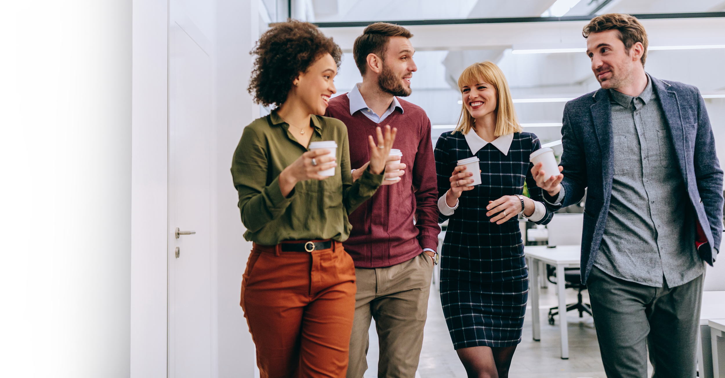 Four coworkers walking and talking together in an office, holding coffee cups, smiling and enjoying their conversation.