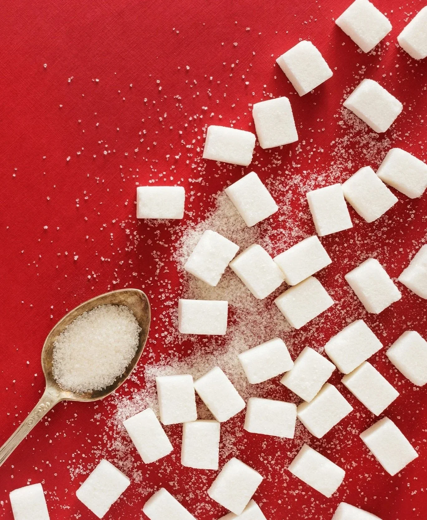 White sugar cubes scattered on a red surface, with a spoon of loose sugar in the lower left corner.