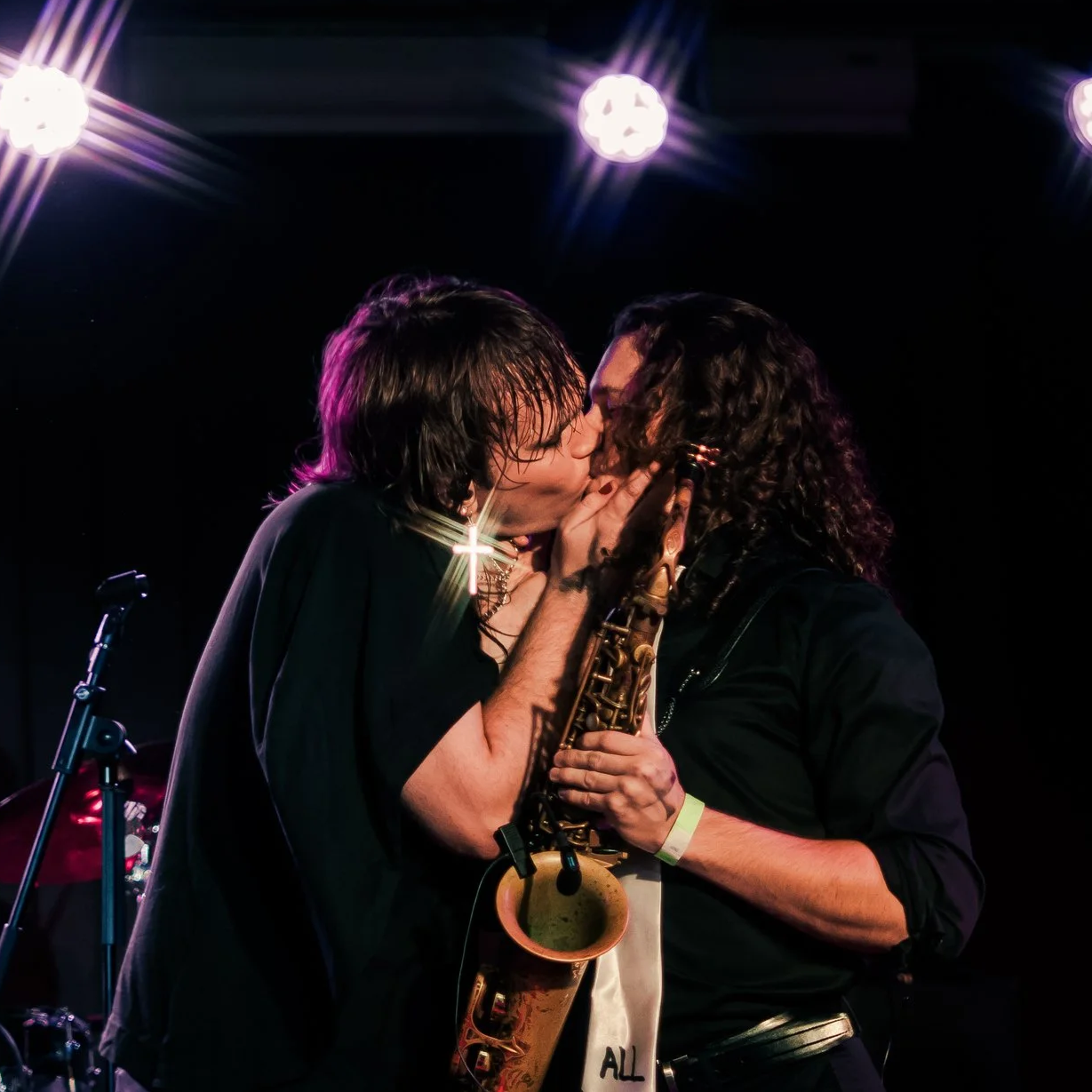Two musicians sharing a kiss on stage, one holding a saxophone, under stage lights.