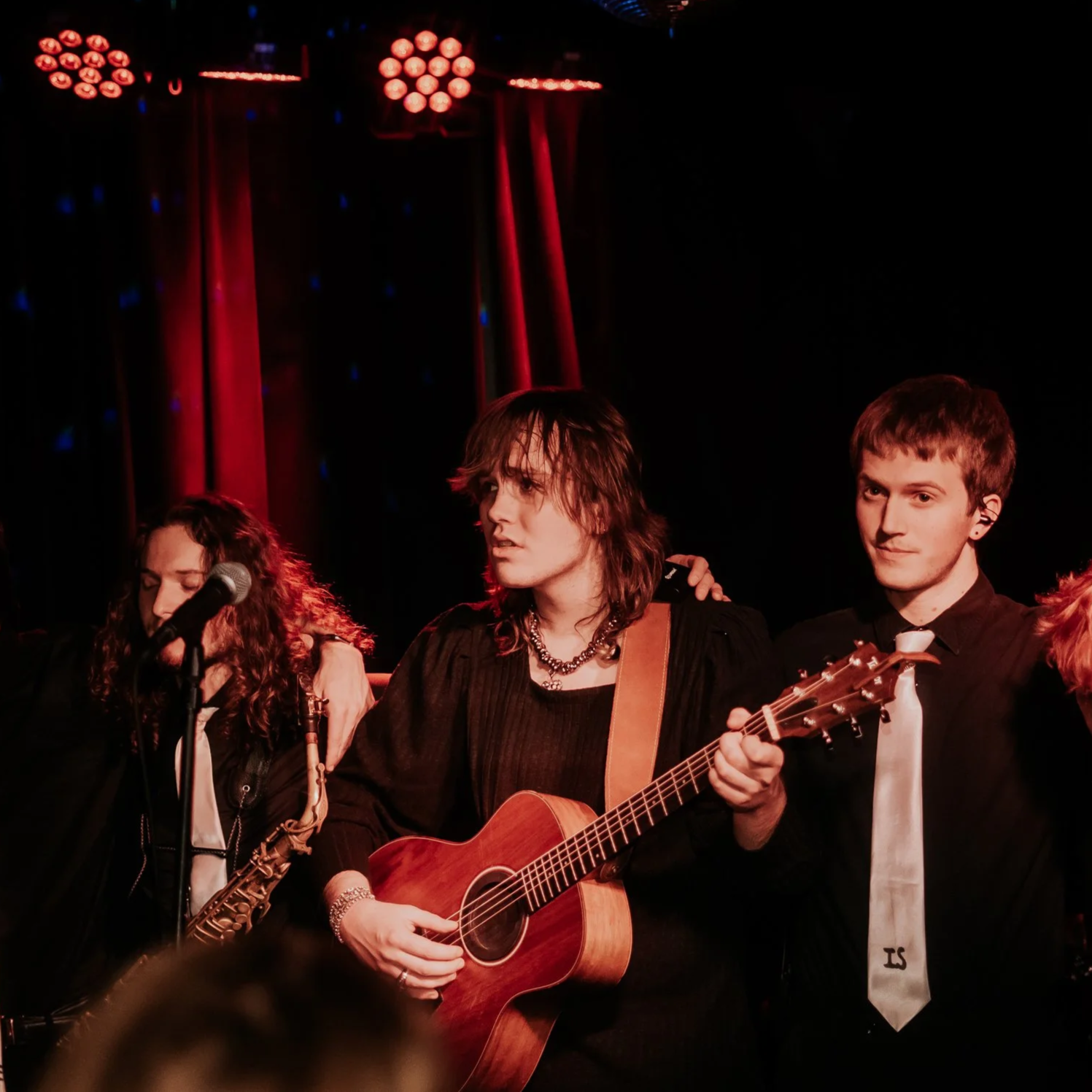 A group of young musicians performing on stage, with one playing an acoustic guitar, a female with curly hair with a microphone and another young man with short hair holding a bass guitar, against a backdrop with red curtains and stage lighting.