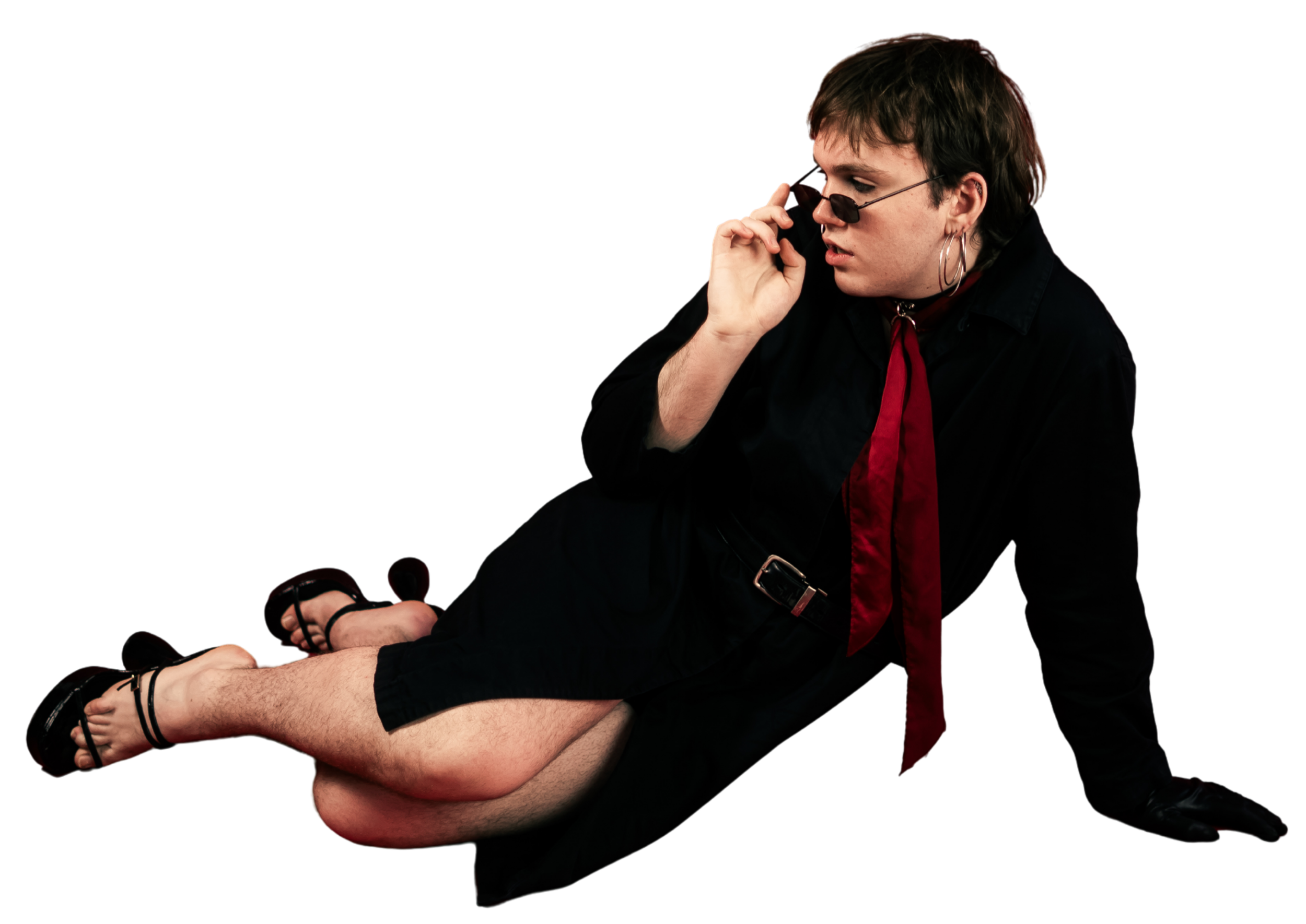 A person with short dark hair, wearing sunglasses and hoop earrings, is wearing black clothing with a red scarf and is sitting close to another person's legs, which are outstretched and barefoot. The scene has a black background.