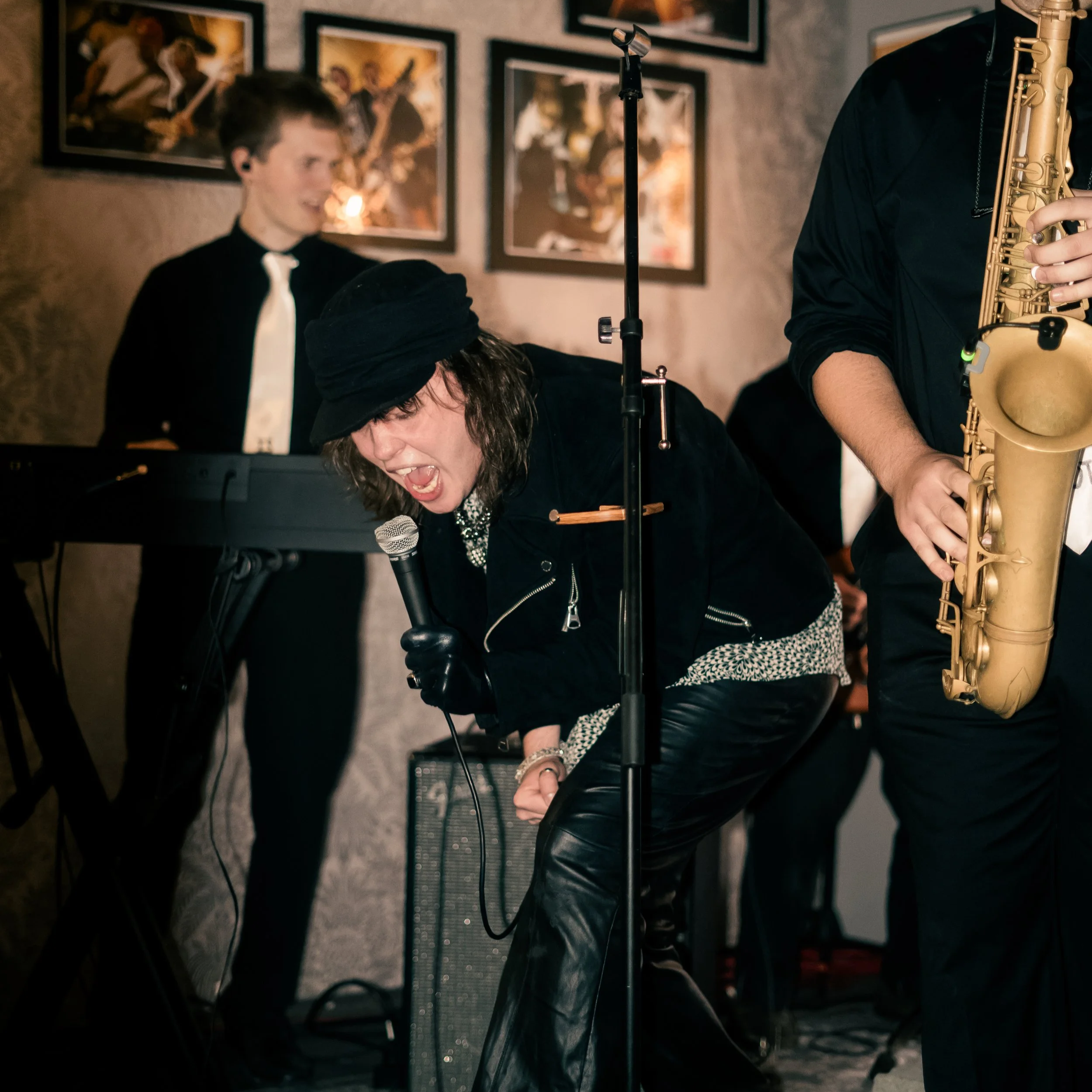 A woman singing into a microphone, a person playing the keyboard, and a person playing the saxophone during a live music performance in a dimly lit venue with framed pictures on the wall.