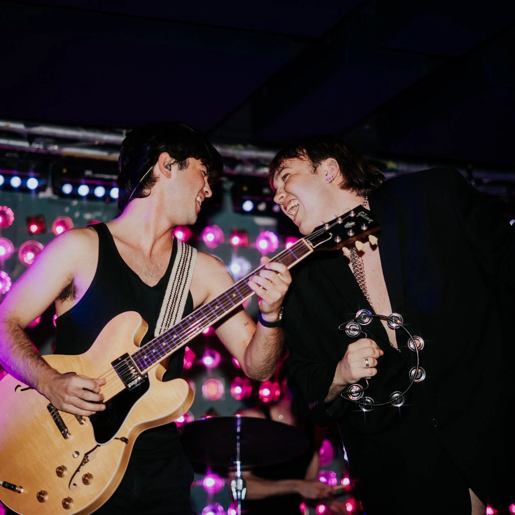 Two men smiling and singing together on stage; one playing an acoustic guitar, the other holding a tambourine; colorful lights in the background.