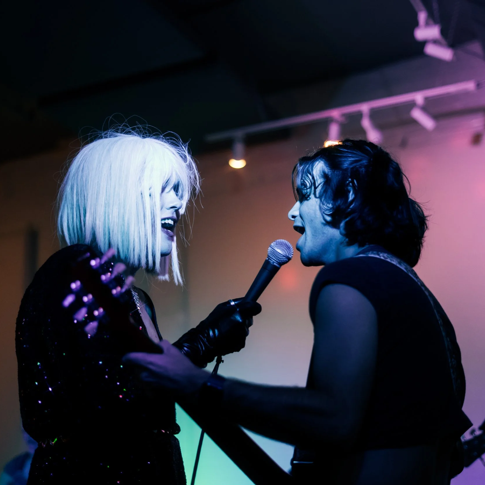 Two performers singing passionately on stage, one with platinum blonde hair and the other with black, holding a microphone, illuminated by colorful stage lighting.
