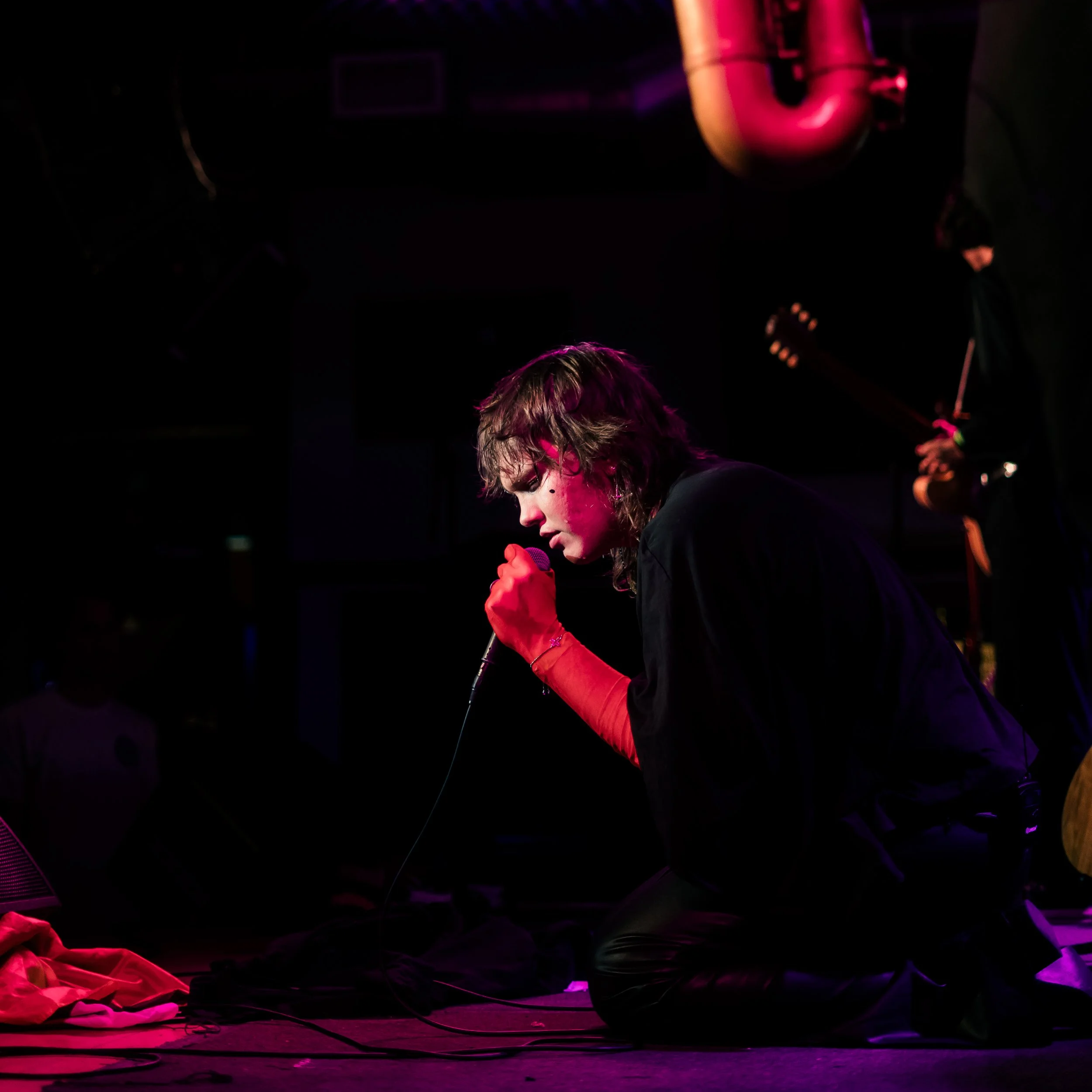 Person with shoulder-length blonde hair and glasses singing passionately into a microphone on stage, with colorful stage lighting and cloth on the floor.