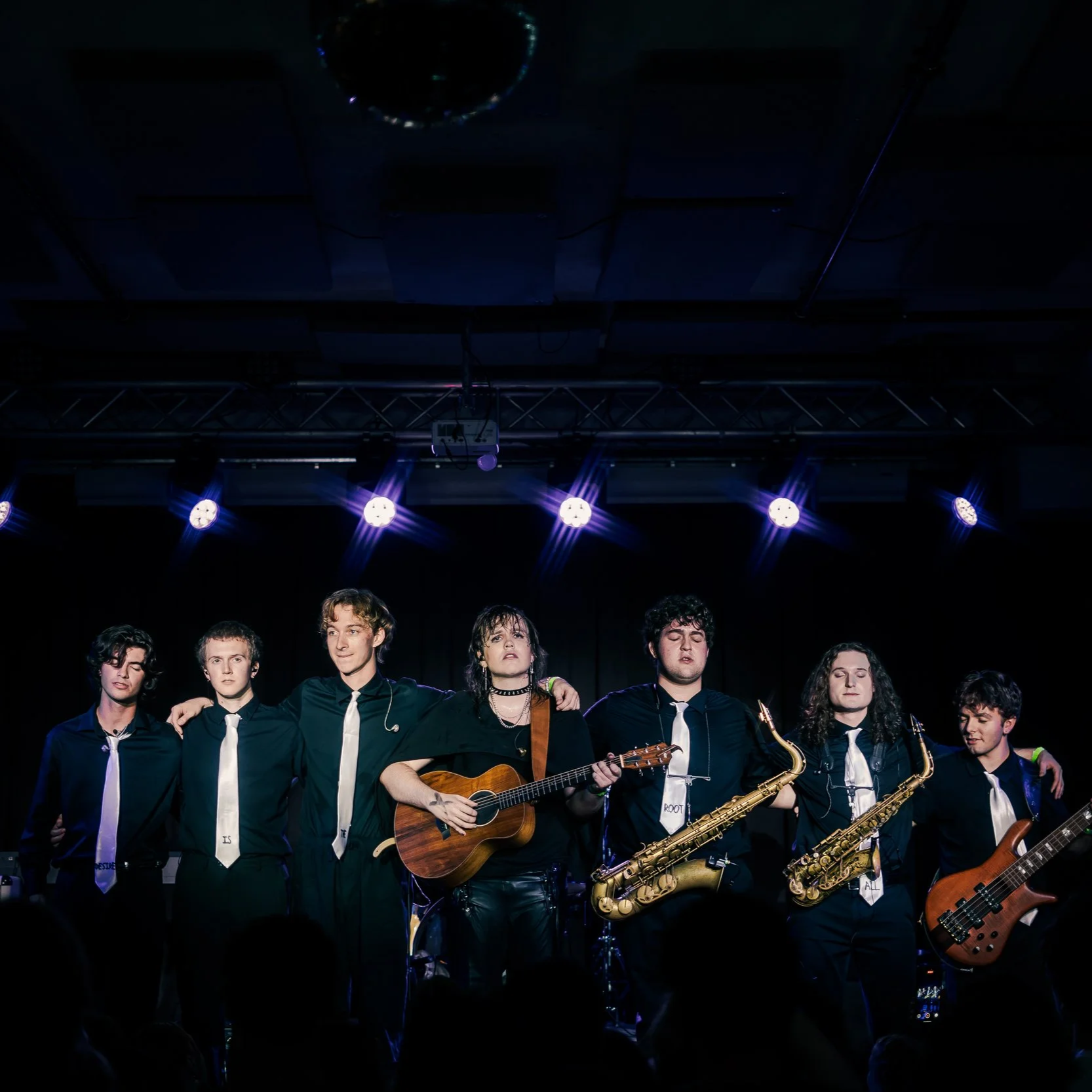 A group of young musicians on stage with various instruments, under stage lighting.