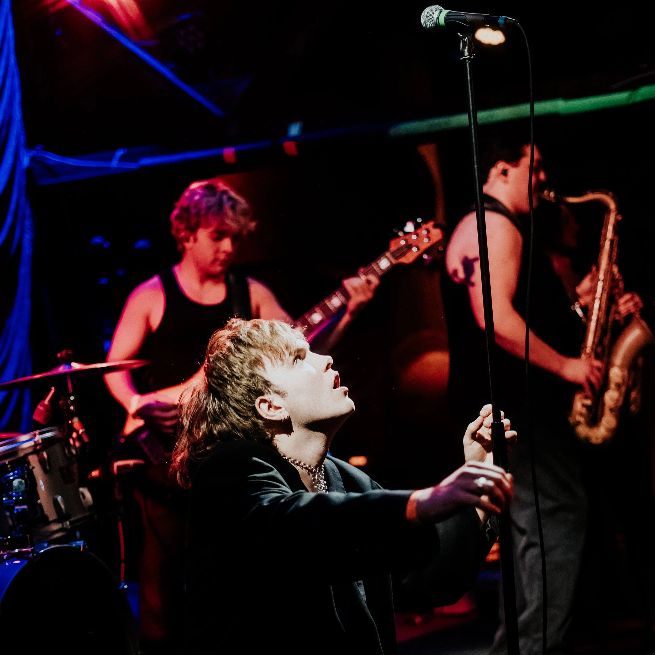 Young man with short blonde hair playing an electric guitar on stage, microphone and other band members visible, colorful stage lighting.