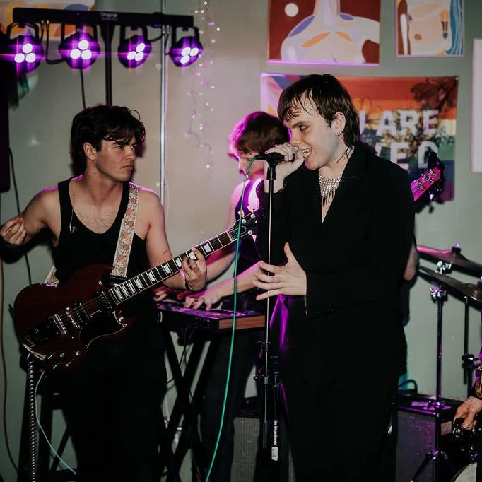 A band performs on stage, with a singer in a black suit singing into a microphone, a guitarist in a tank top playing electric guitar, and a keyboardist in the background. Colorful stage lighting and posters are visible.