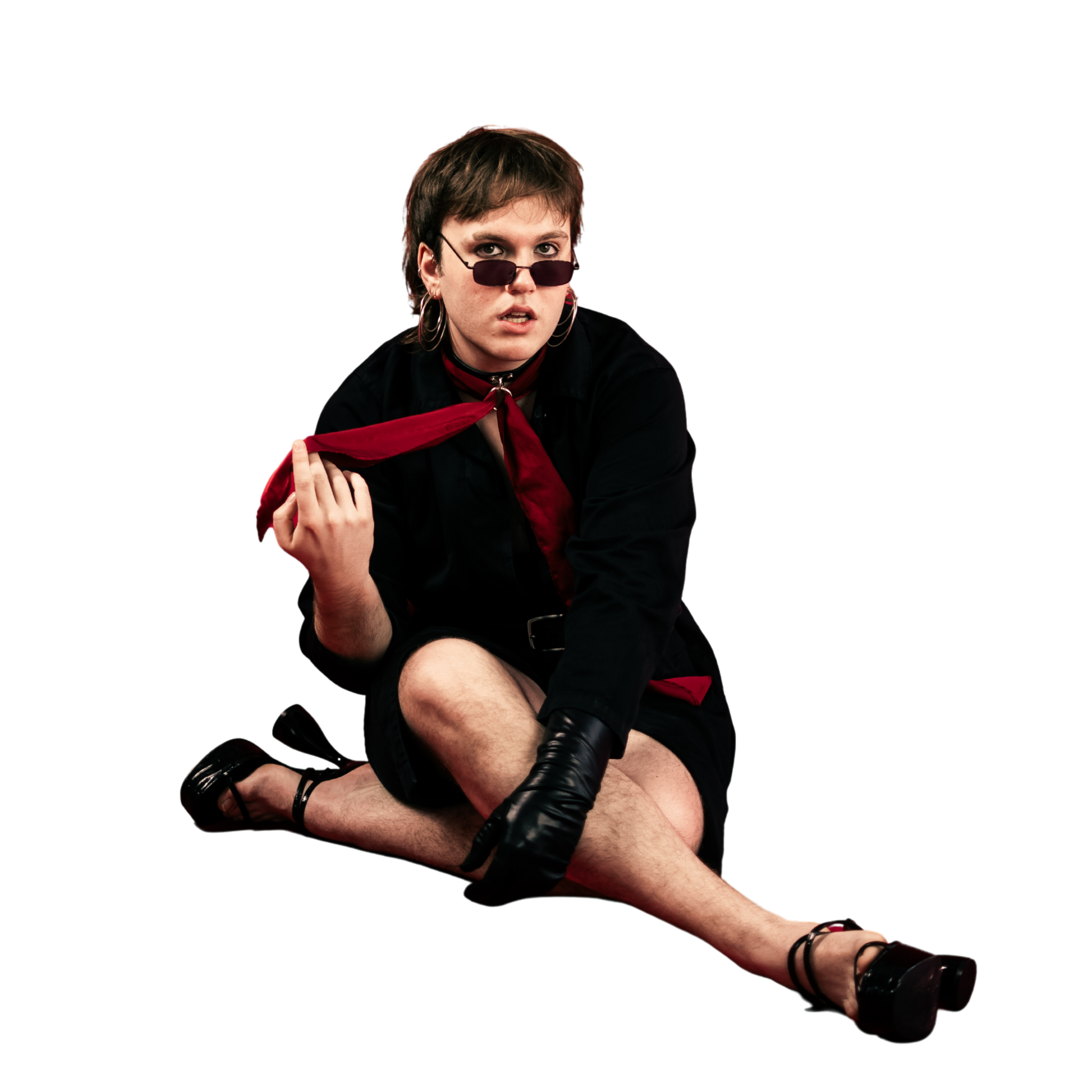 A person with short brown hair wearing black sunglasses, hoop earrings, a red neck scarf, and black clothing, sitting with crossed arms, against a black background.