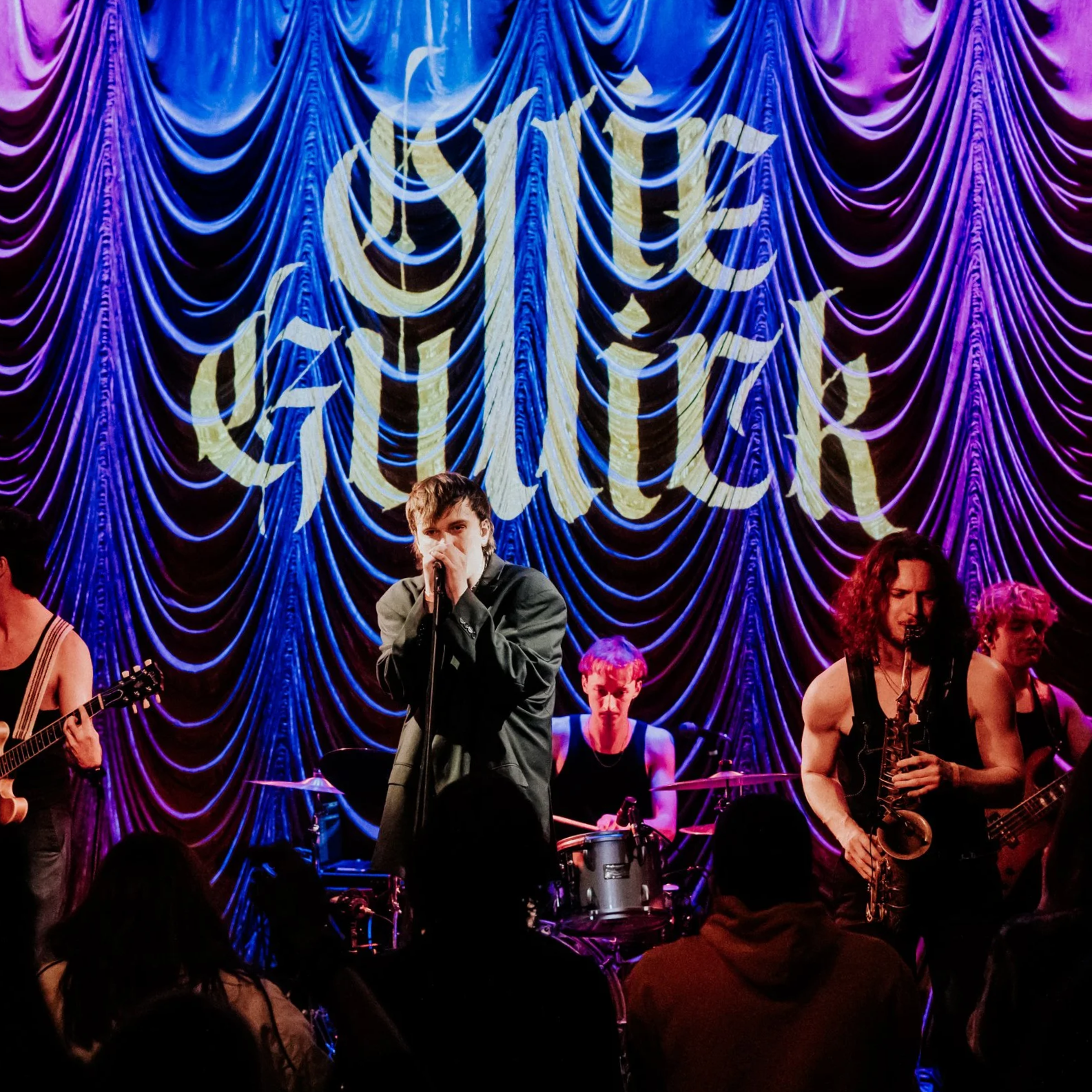 A band performs on stage with a purple curtain backdrop and the words 'Live music' illuminated in gold behind them. The band members include a singer, guitarists, a drummer, and a saxophonist in a small music venue.
